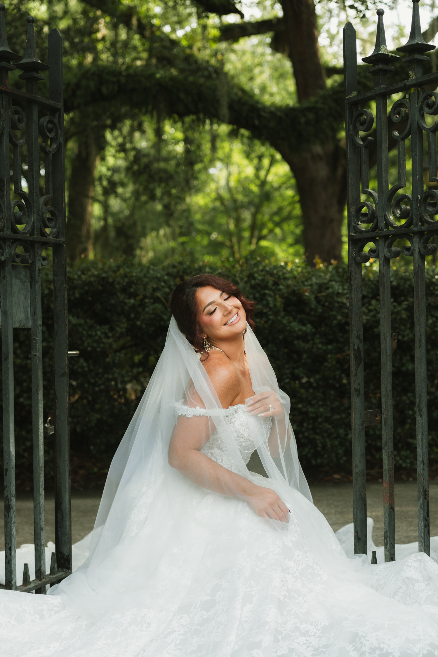 Smiling bride in white ball gown and veil sits gracefully before ornate iron gates surrounded by lush greenery.