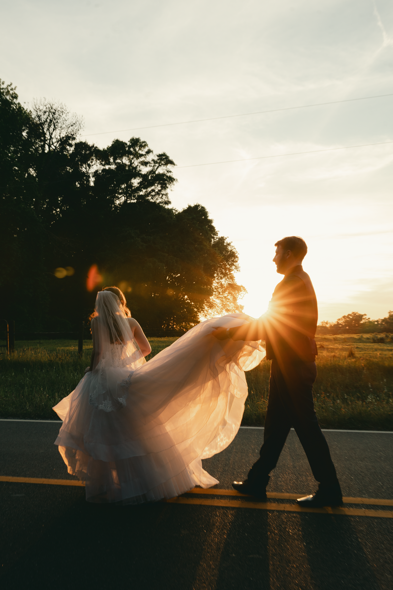 Bride and groom walking on a road at golden sunset, wedding dress flowing in the wind with sun flare behind them.