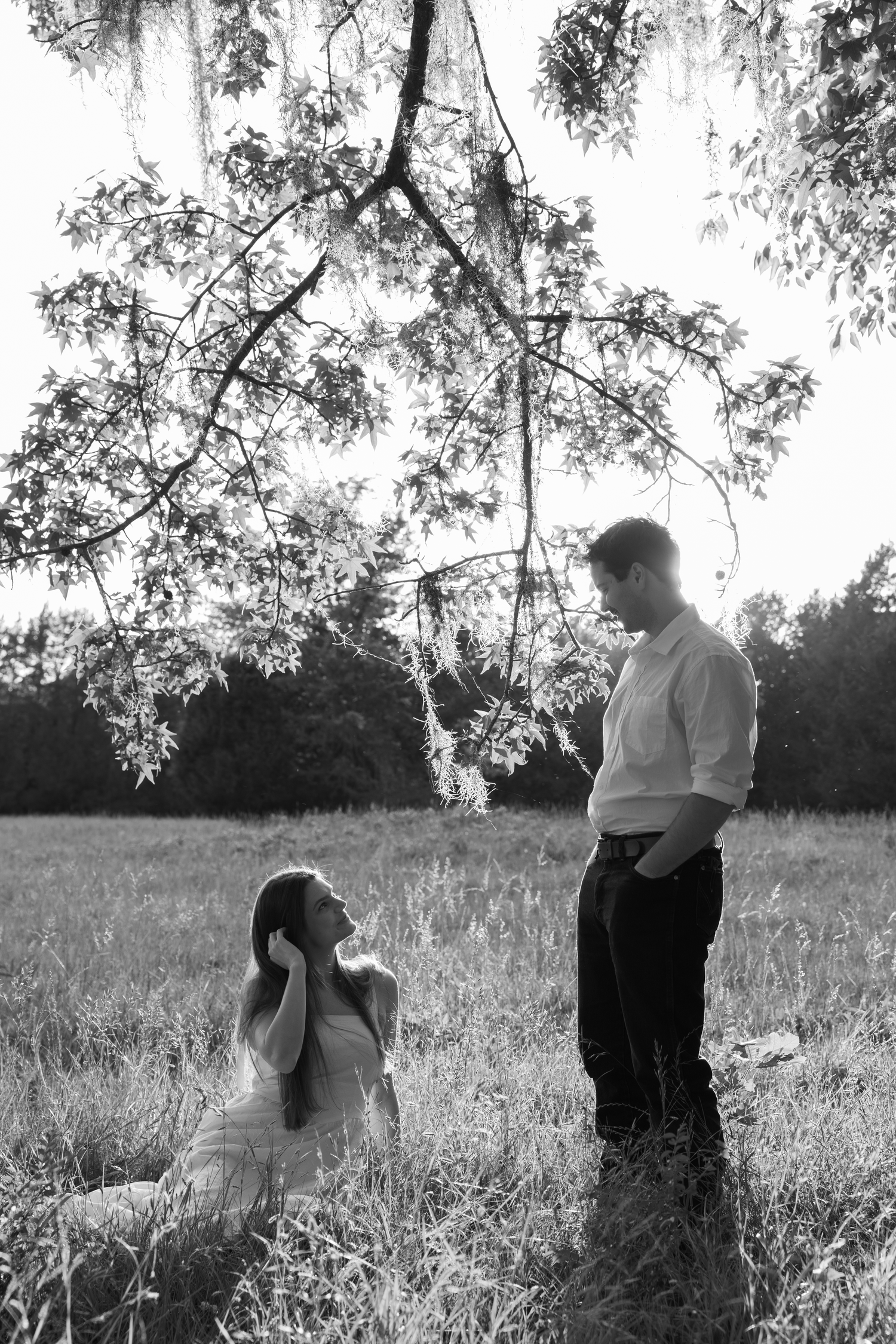 Black and white photo of a couple in a field, woman sitting in grass looking up at standing man under tree branches.