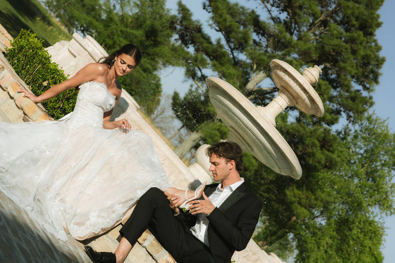 Bride in white gown and groom in tuxedo sitting by ornate stone fountain outdoors on their wedding day.
