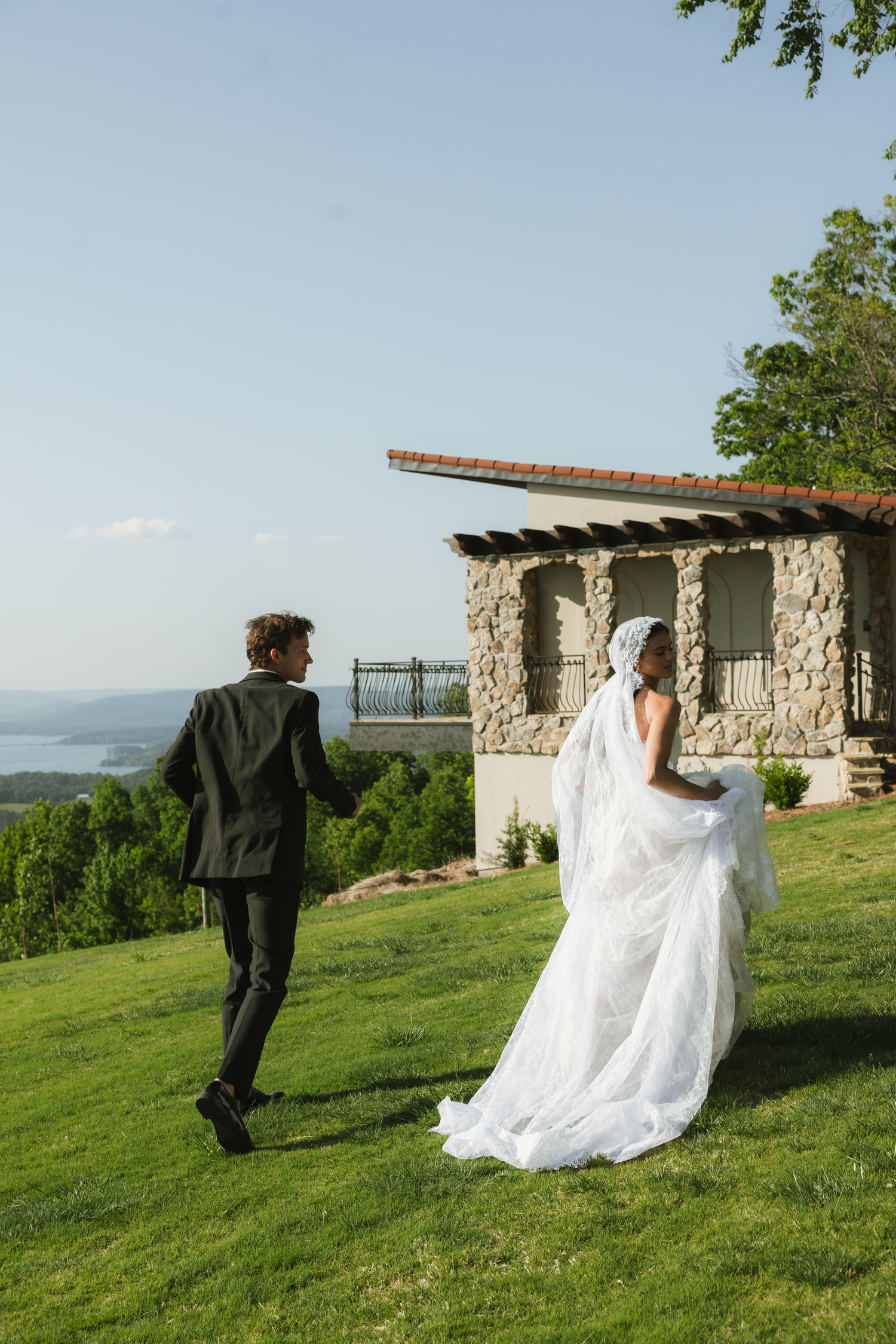 A bride in a flowing white gown and veil walks with a groom in a dark suit on a lush green hillside venue.