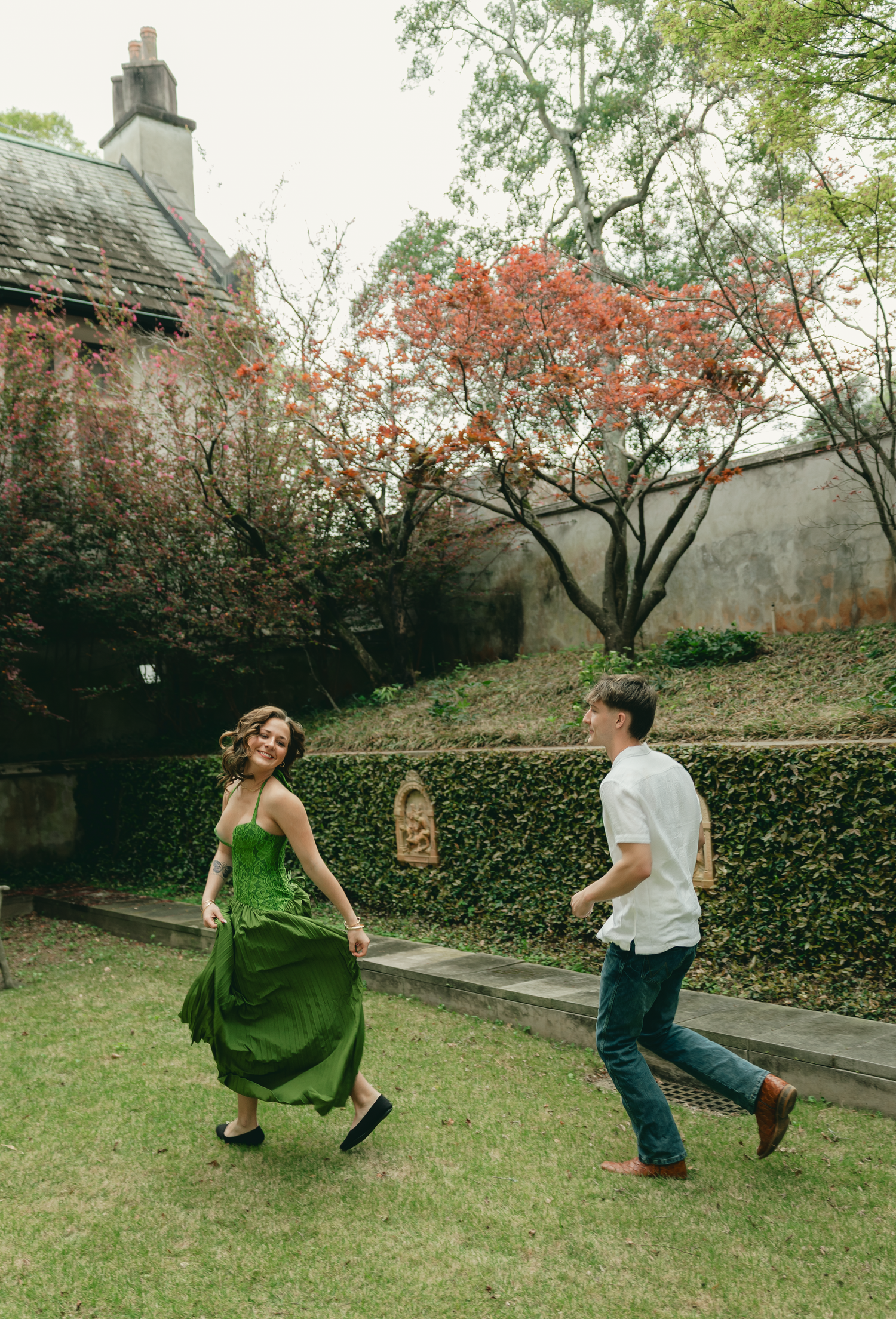 Couple joyfully running through a lush garden, woman in green dress, man in white shirt and jeans, red flowering tree backdrop.