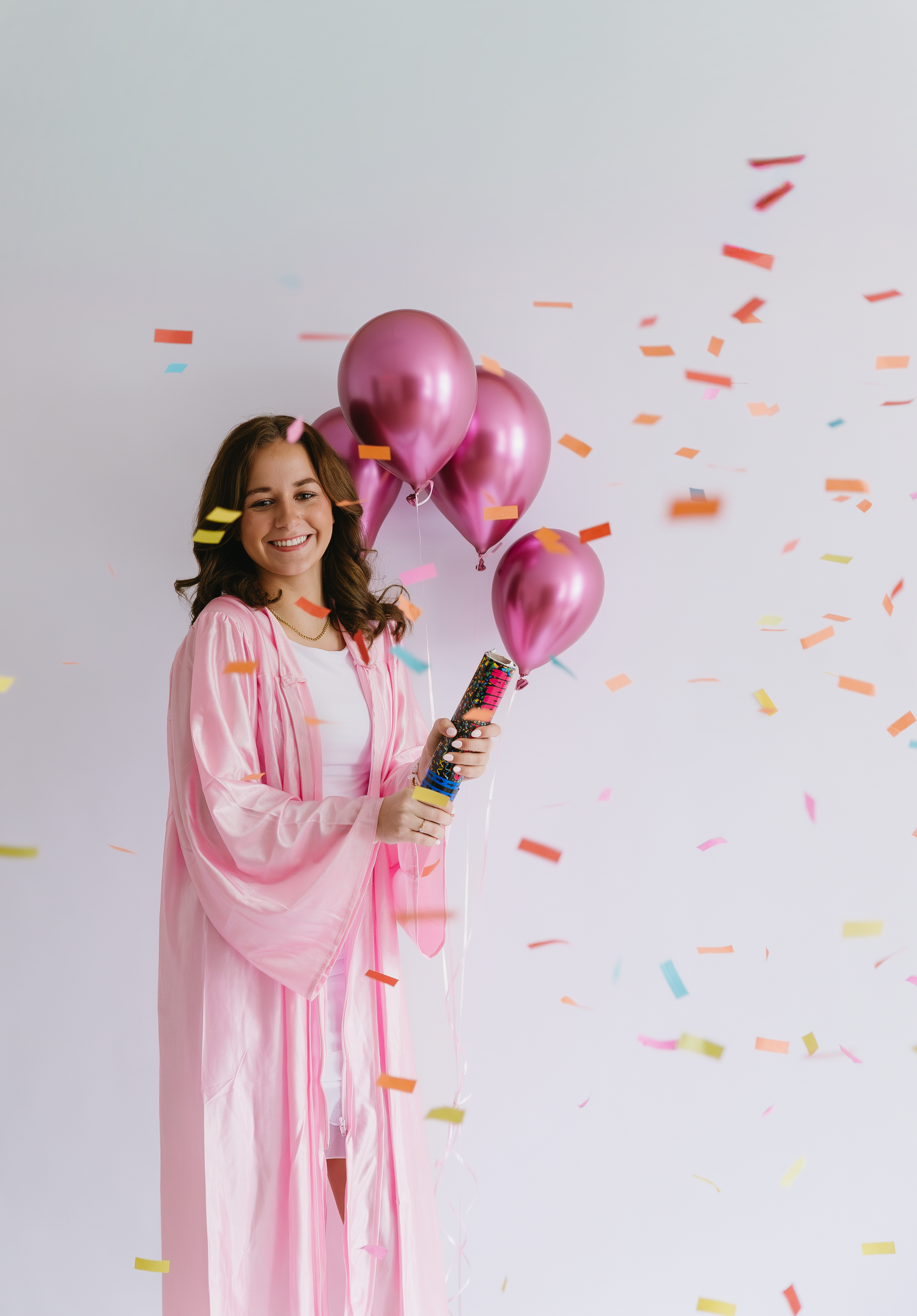 Woman in pink robe holding magenta balloons and confetti cannon, celebrating with colorful confetti falling around her for her senior photos