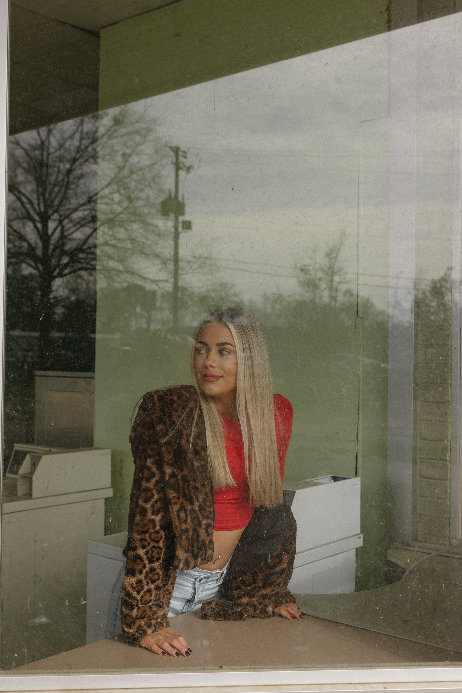 Young woman in leopard print jacket and red crop top sits behind a glass window, gazing thoughtfully to the side.