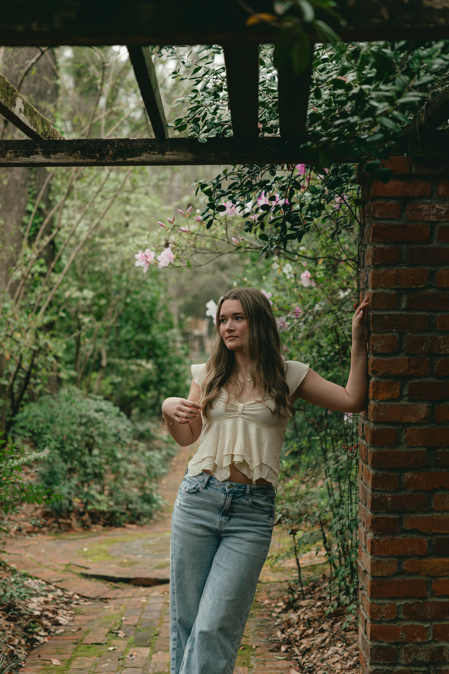 Young woman in jeans and cream top poses by brick wall in lush garden with pink blooming flowers.