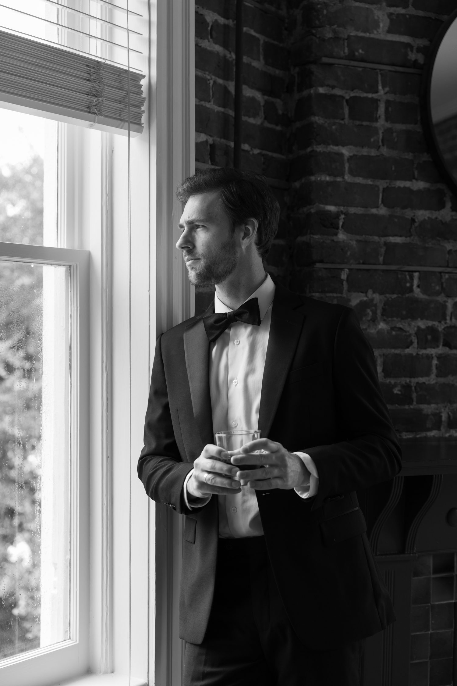 Black and white photo of a groomsman in a tuxedo holding a whiskey glass while gazing out a window.