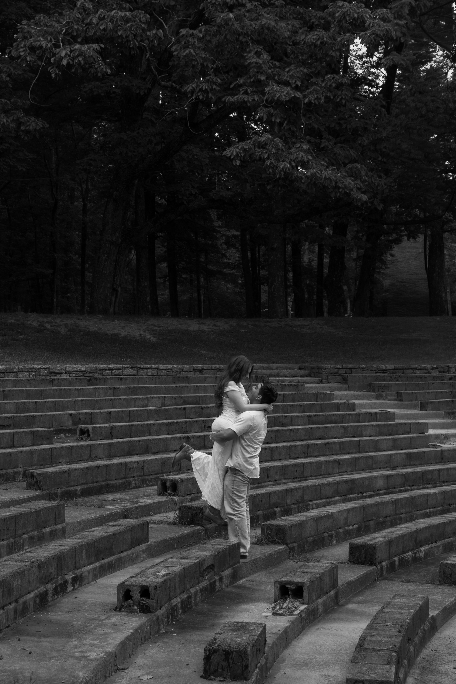 Black and white photo of a couple embracing on stone amphitheater steps, surrounded by tall trees.