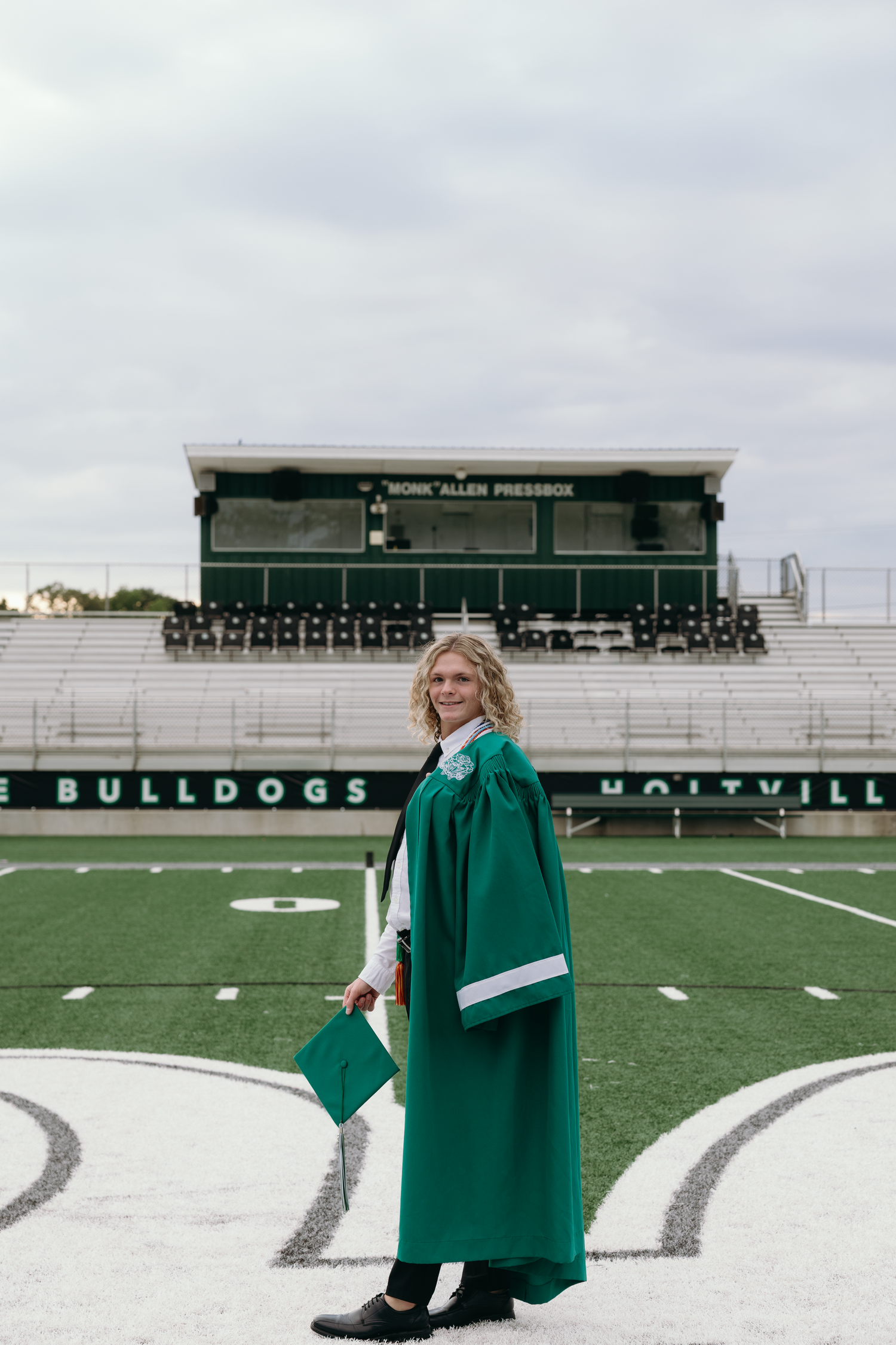 A smiling graduate in green cap and gown stands on the Holtville Bulldogs football field.
