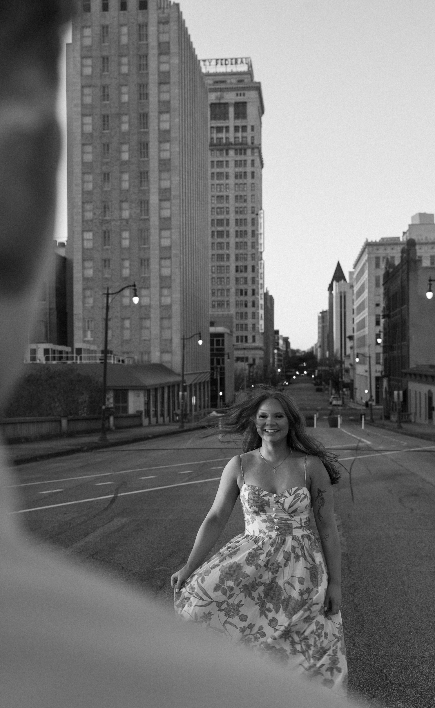 Black and white photo of a smiling woman in a floral dress walking on an empty urban street with tall buildings.