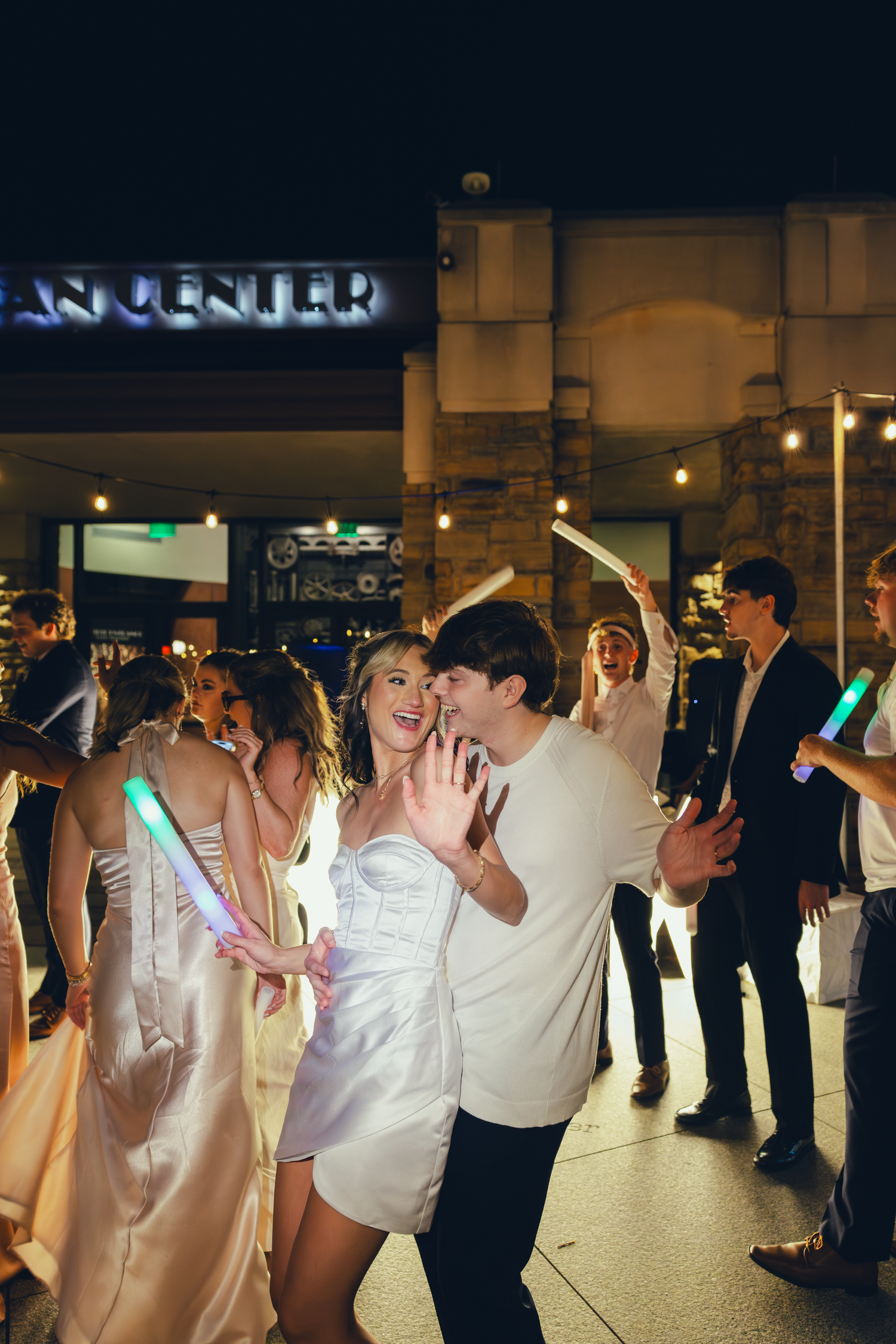 A joyful couple dances outdoors at night holding glowing light sticks surrounded by wedding guests near a lit venue.