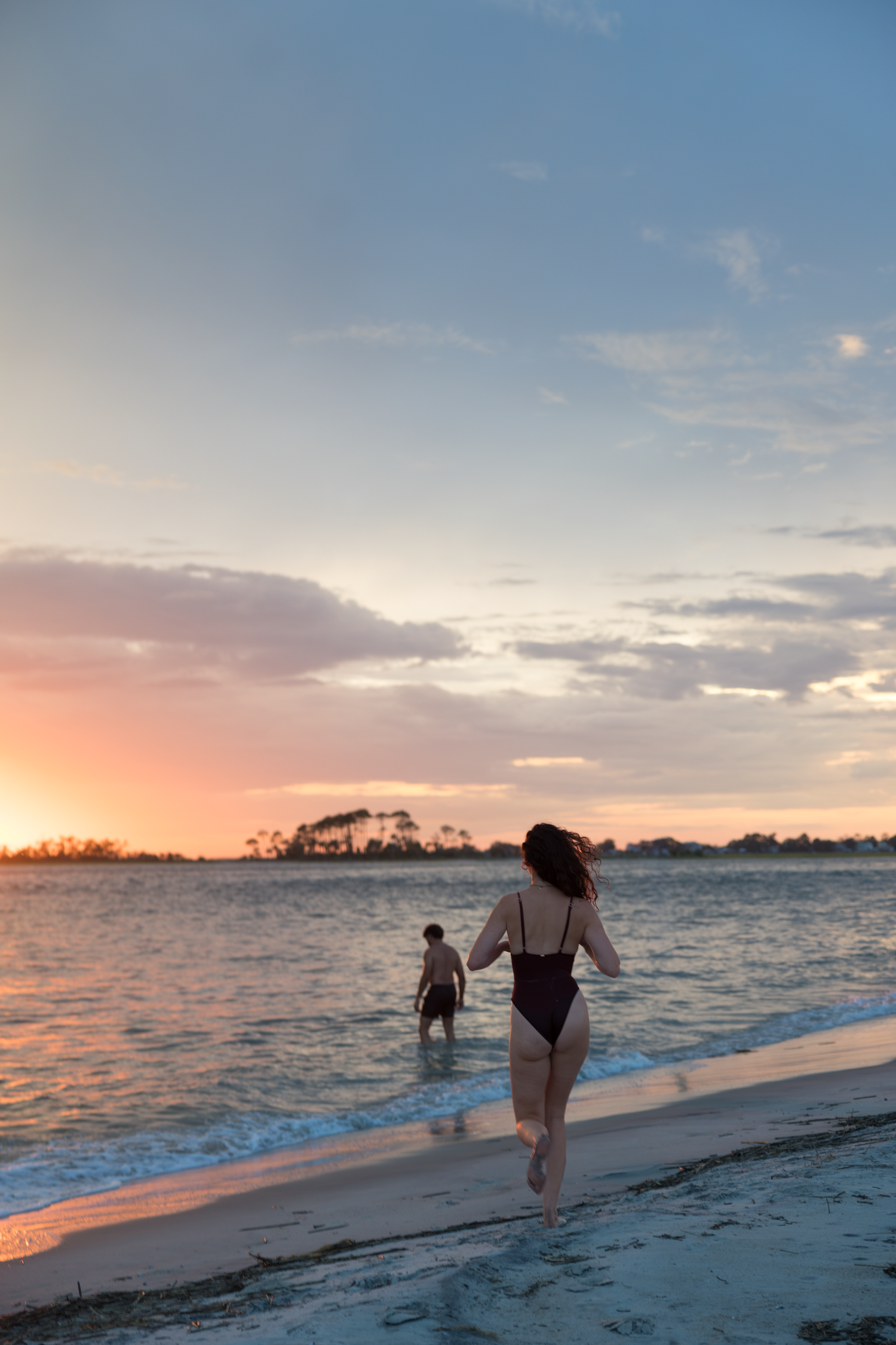 Two women in swimsuits walk toward ocean waves at a stunning tropical beach sunset with colorful sky.