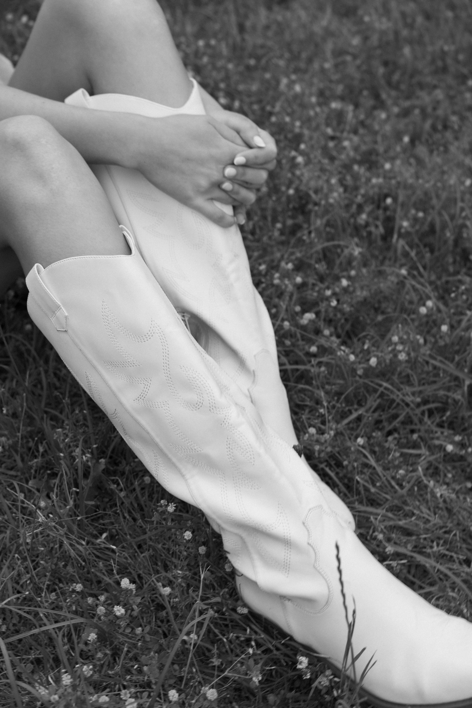 Black and white photo of a woman sitting on grass wearing white cowboy boots and a white dress.