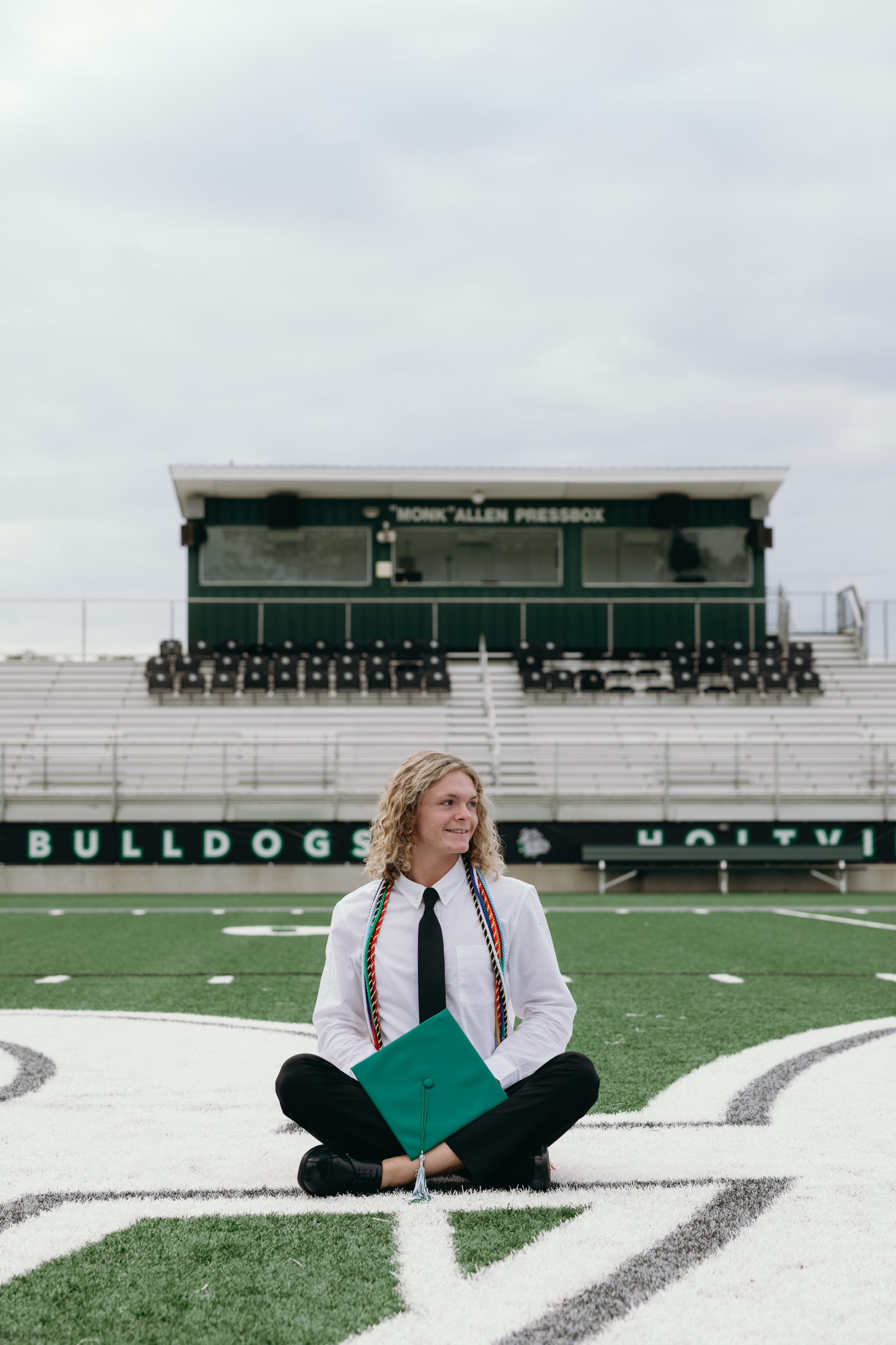 Graduate sits cross-legged on Bulldogs football field holding green cap, wearing white shirt and tie.