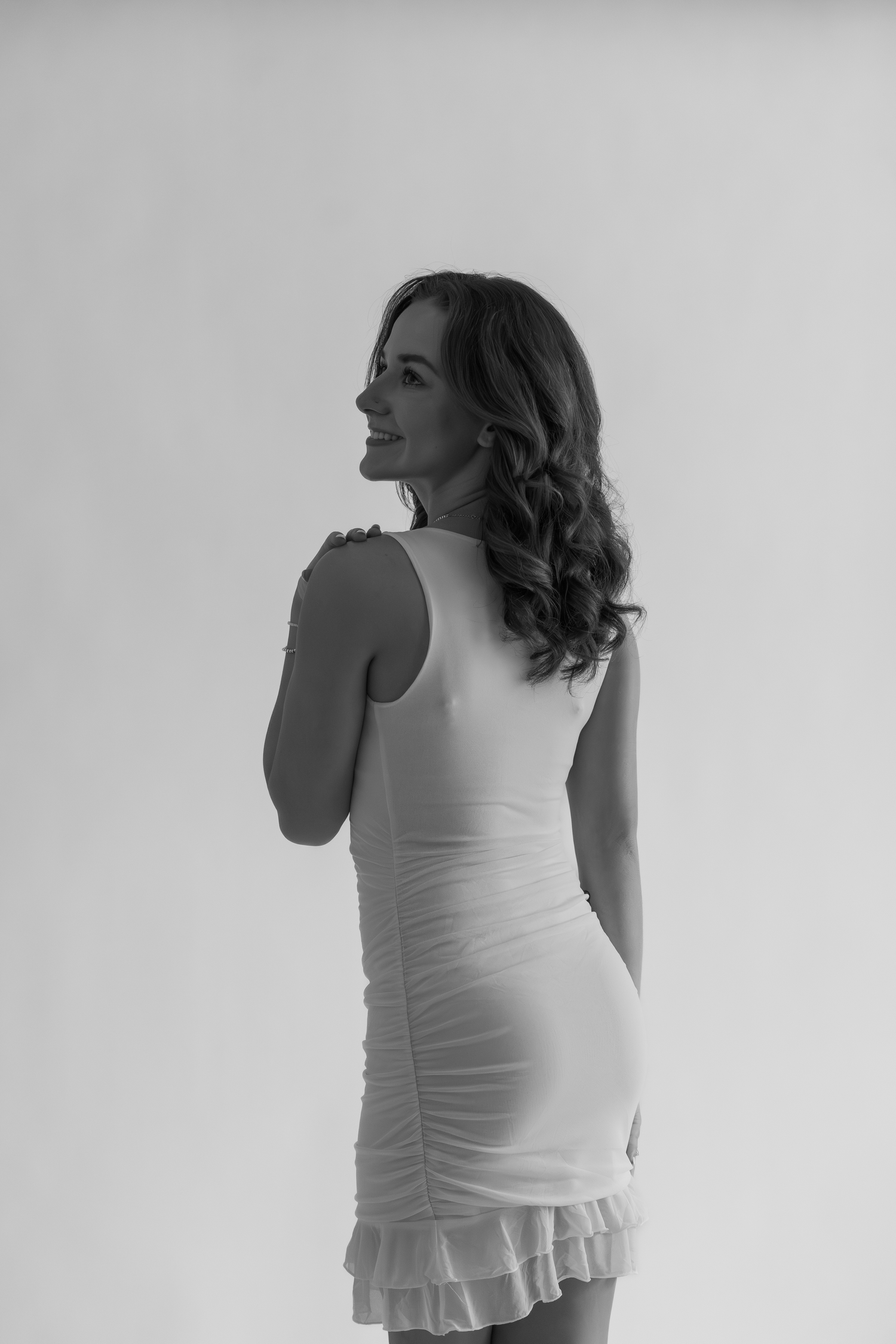 Black and white photo of a woman in a fitted white ruffle dress, posing with her back turned, smiling over her shoulder.