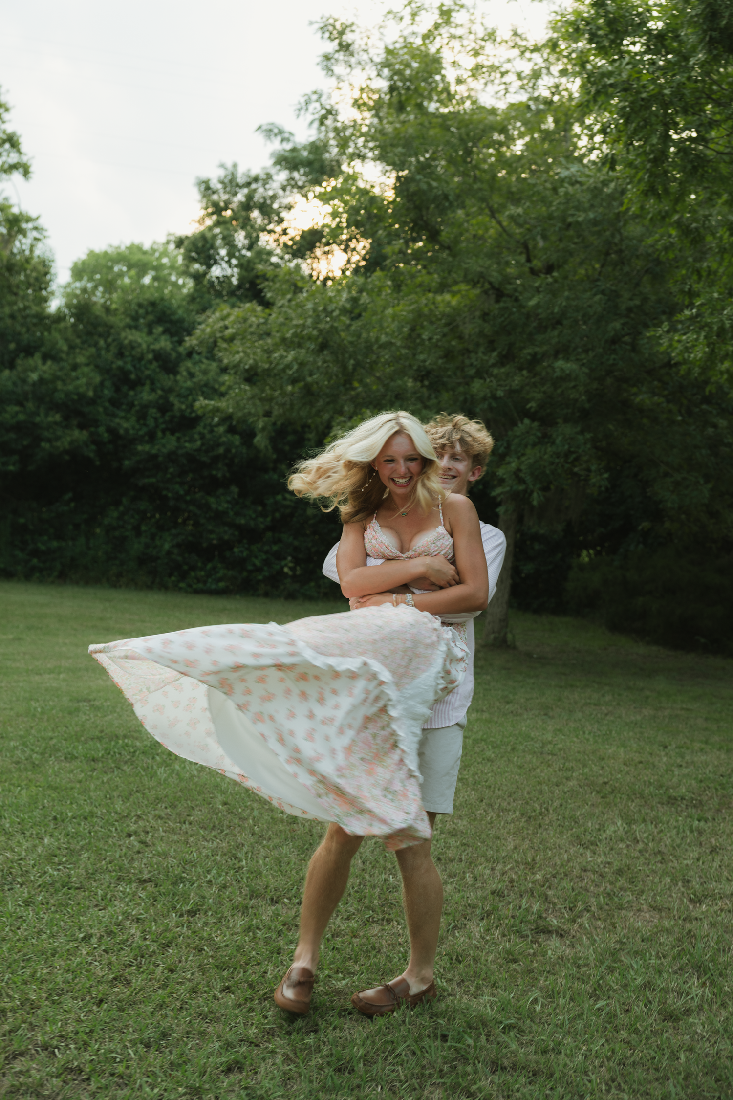 A joyful couple dancing outdoors, man hugging woman in floral dress with flowing hair, surrounded by lush green trees.