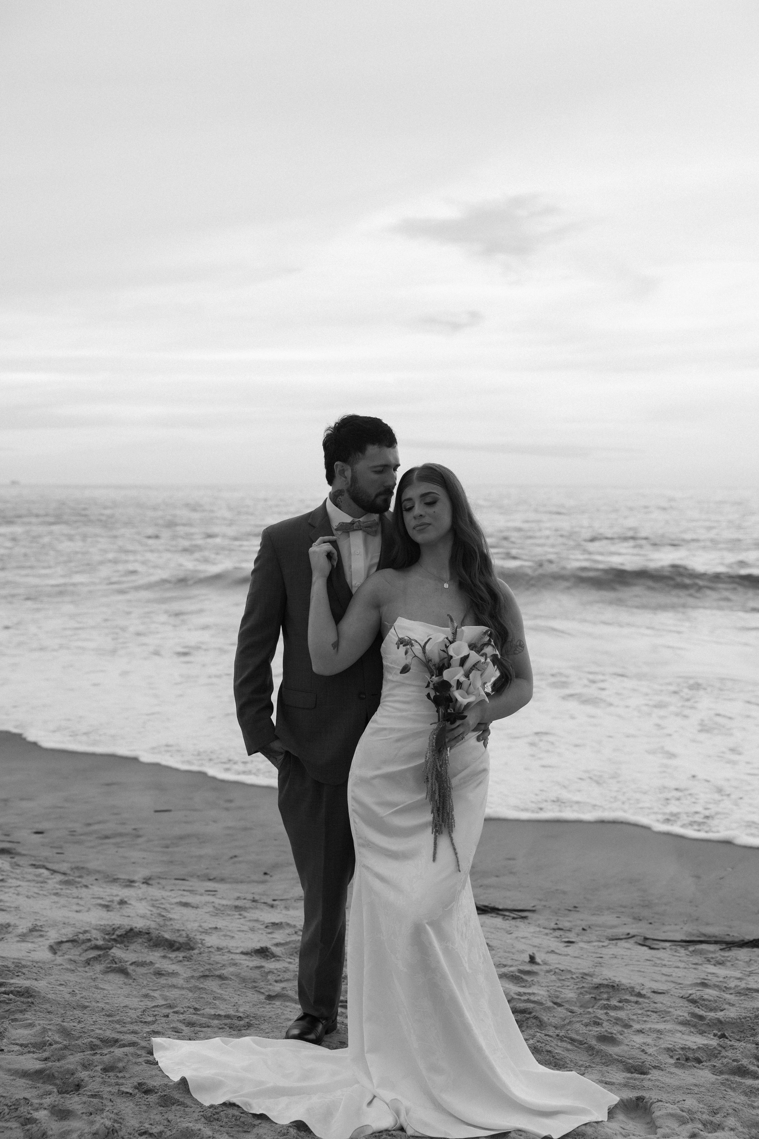 Black and white beach wedding photo of bride holding bouquet and groom in suit embracing by ocean waves.