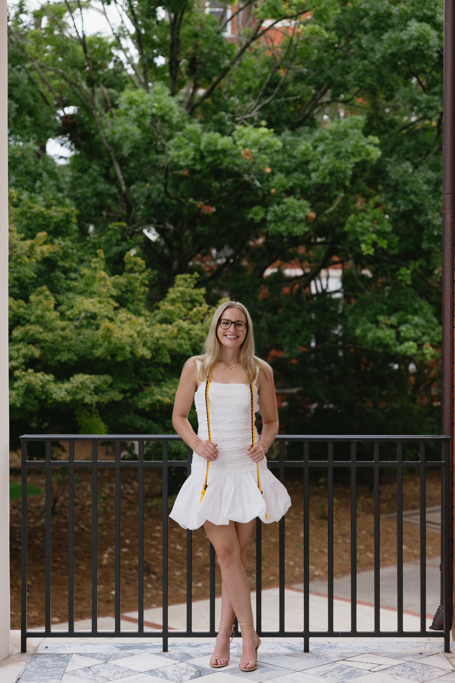 Young blonde woman in white dress and glasses stands on outdoor balcony surrounded by lush green trees.