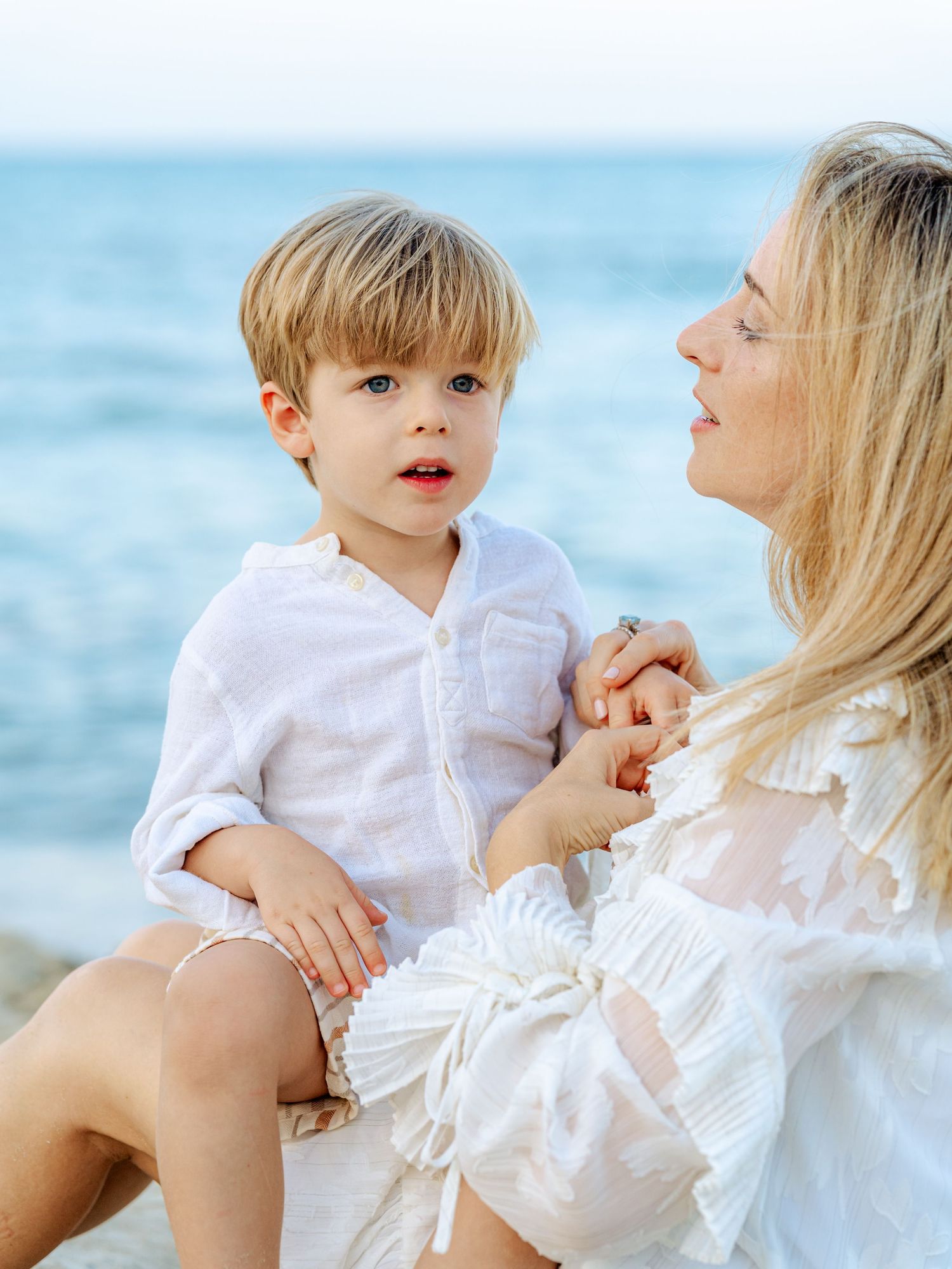 Young blonde boy in white shirt being held by his smiling mother near the sparkling blue ocean.