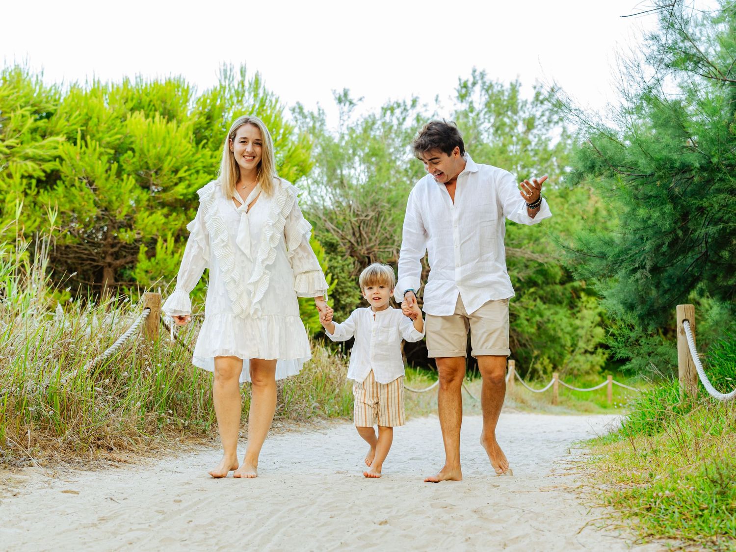 A happy family of three walks barefoot hand-in-hand along a sunny sandy beach path.