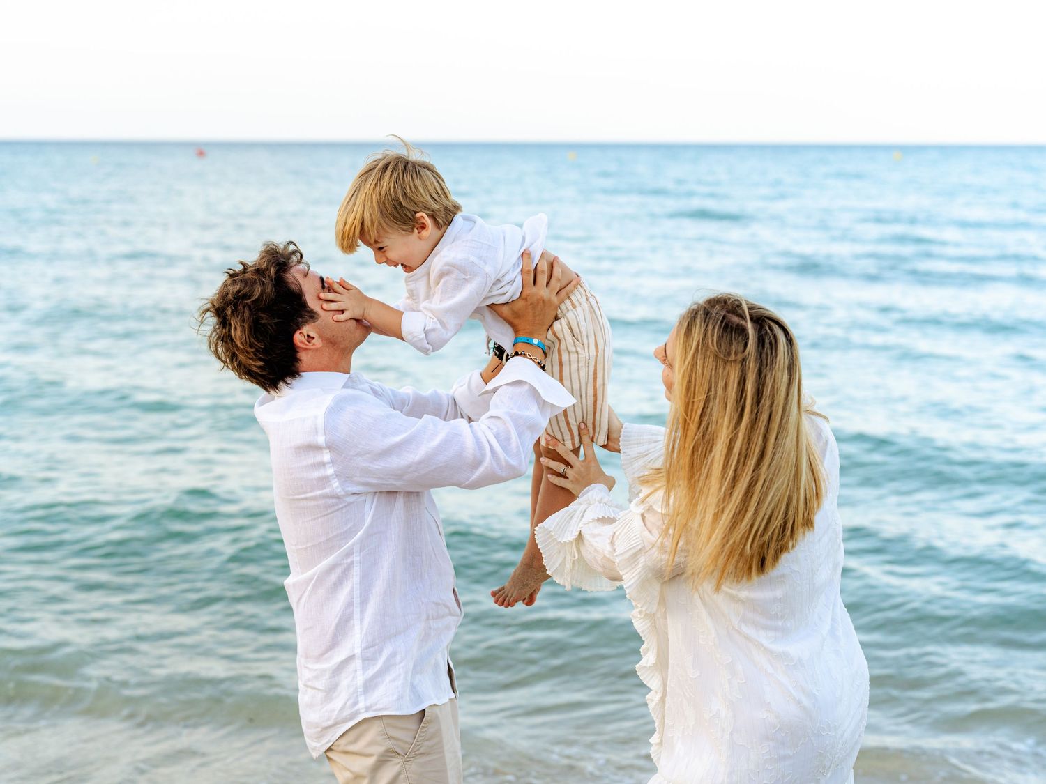 Parents kissing toddler boy on beach with blue ocean backdrop.