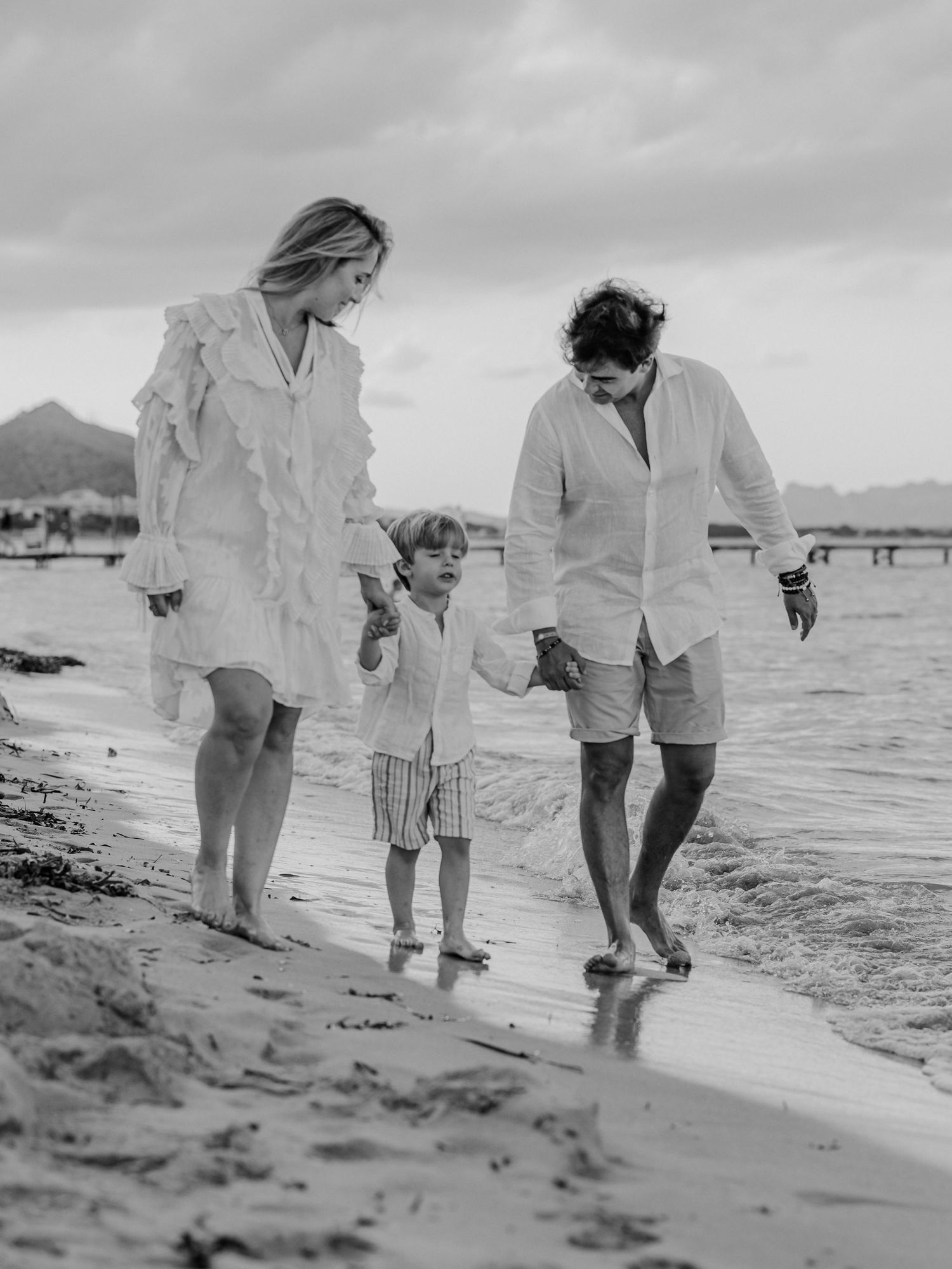 Black and white photo of a family of three holding hands and walking along a beach shoreline.