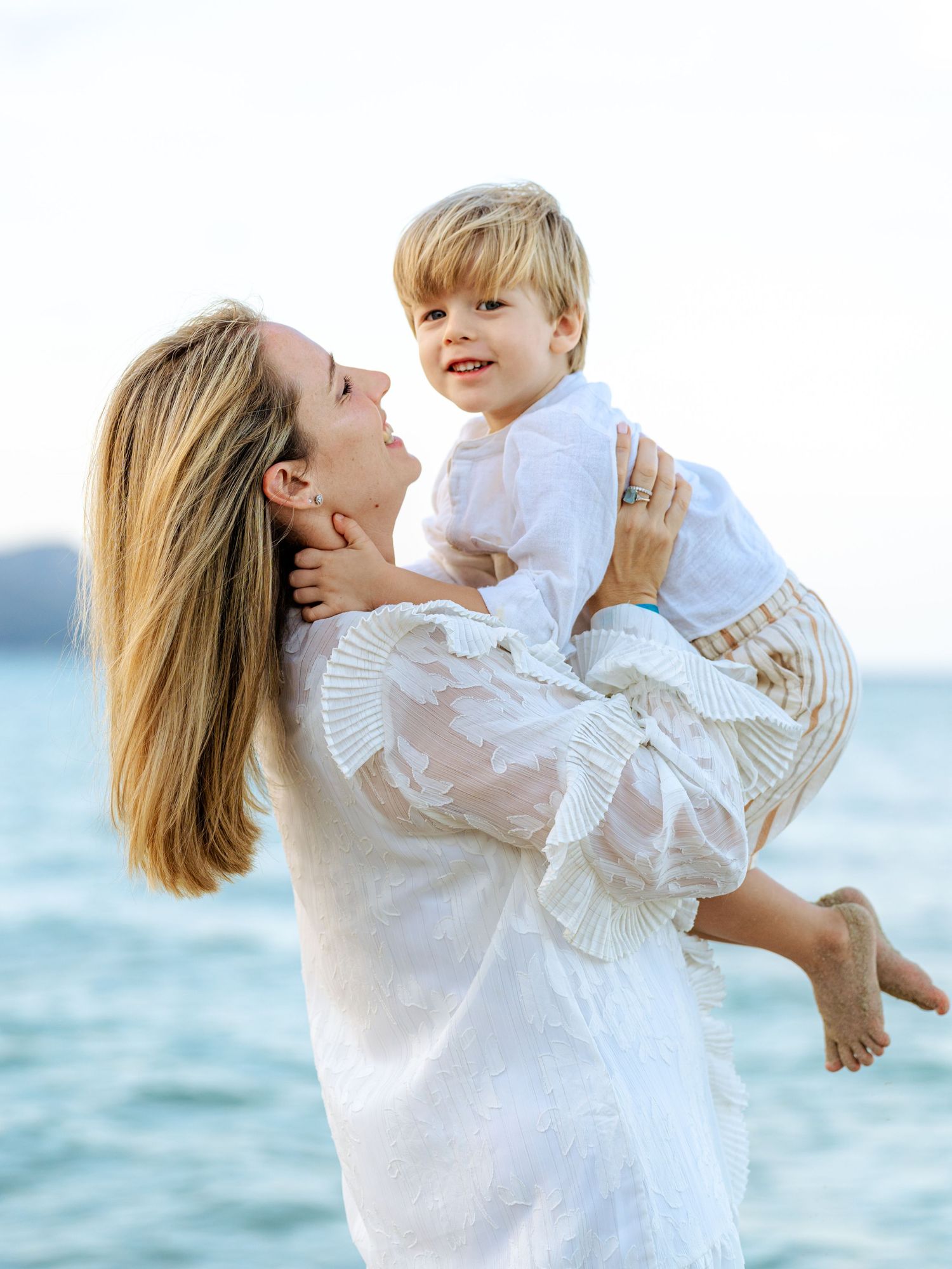 A smiling mother in a white dress lifting her happy blonde toddler boy at the beach.