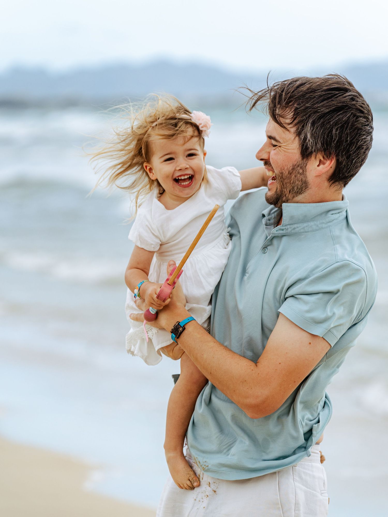 Father in blue shirt laughing while holding happy toddler at beach.