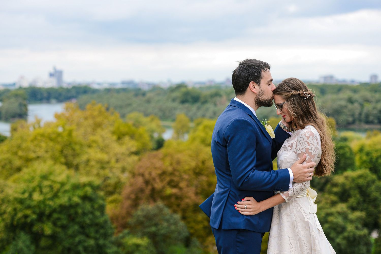 Bride and groom in blue suit embracing with lush green cityscape and river visible in background.