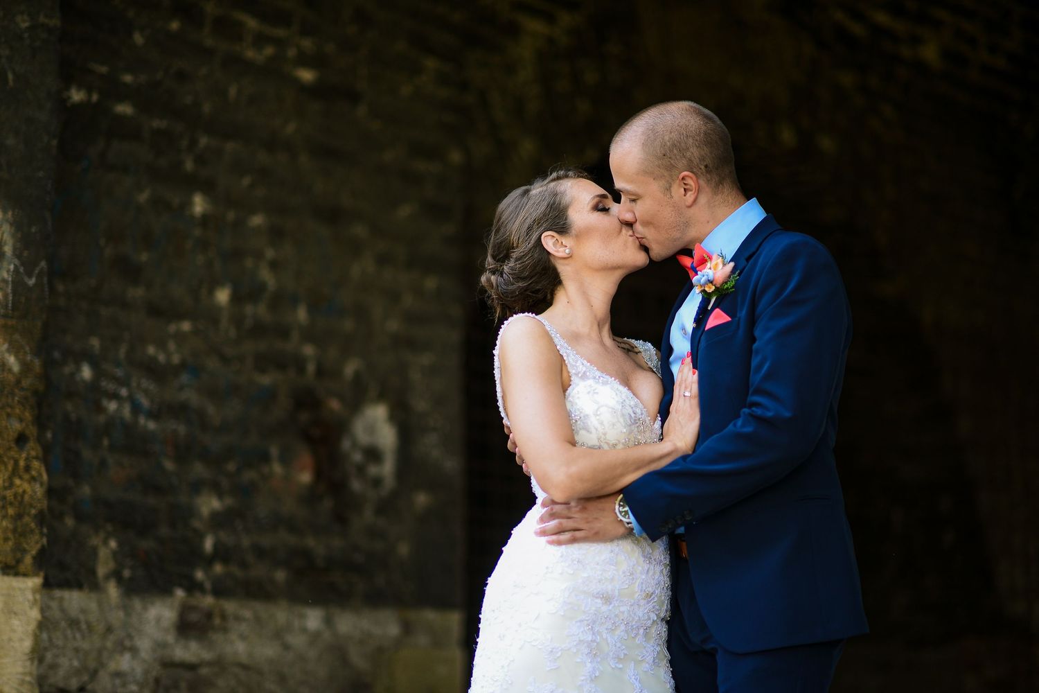 Bride and groom share a kiss in front of a rustic stone wall in their wedding attire.