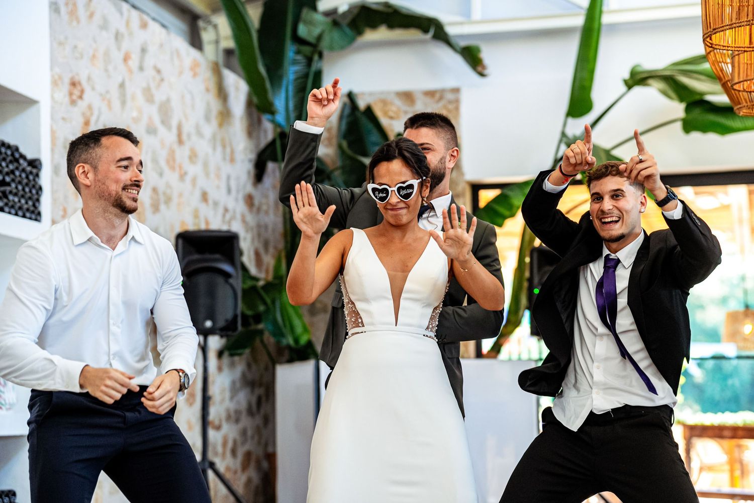 Bride wearing white heart sunglasses dances energetically with guests at a lively wedding reception celebration.