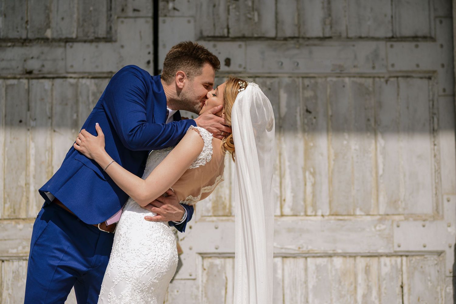 Groom in blue suit embracing and kissing bride in lace wedding dress against a rustic barn wall.
