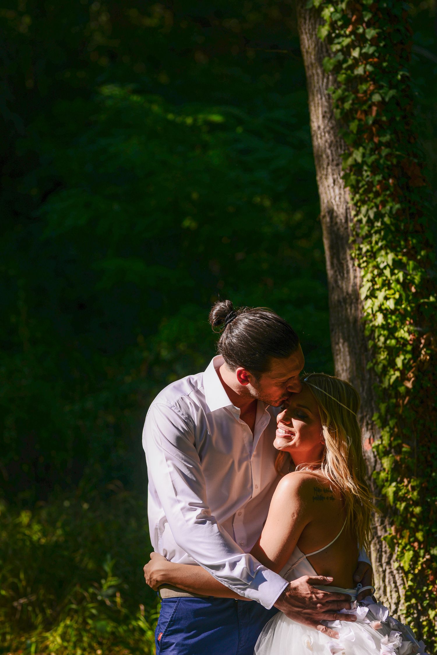 Groom kissing smiling bride beside an ivy-covered tree in a sunlit forest wedding portrait.