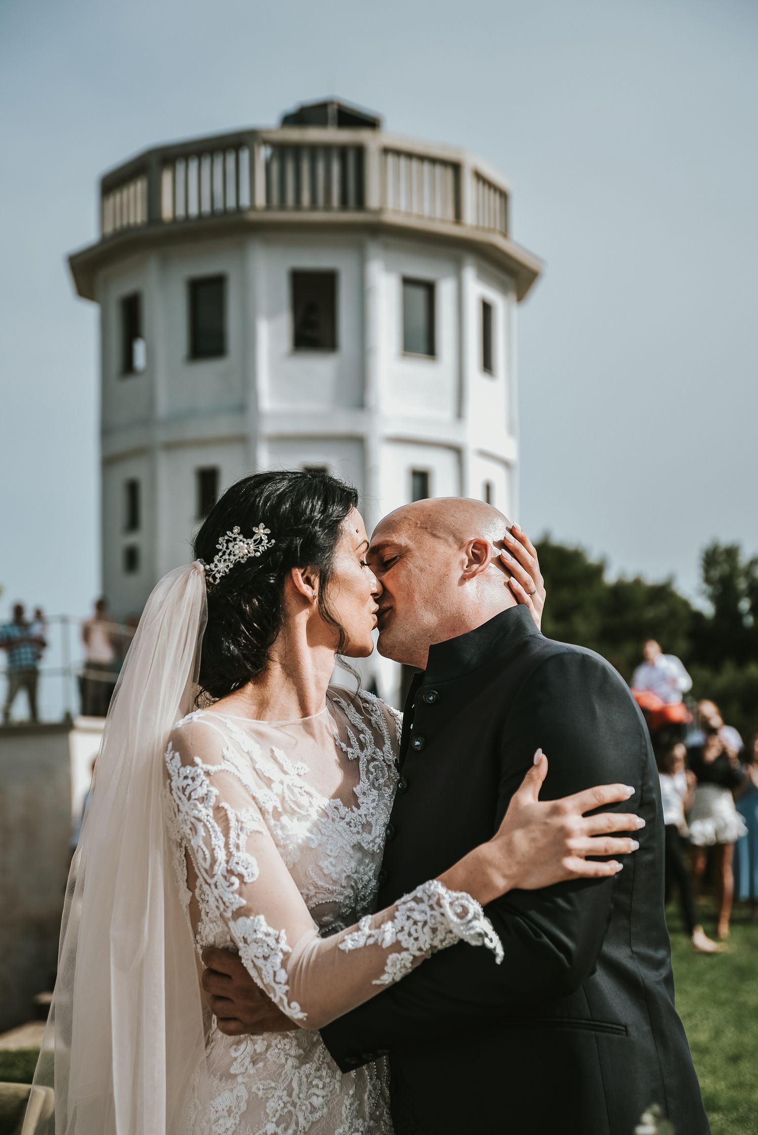 Bride and groom sharing a romantic first kiss with a white tower landmark in the background.