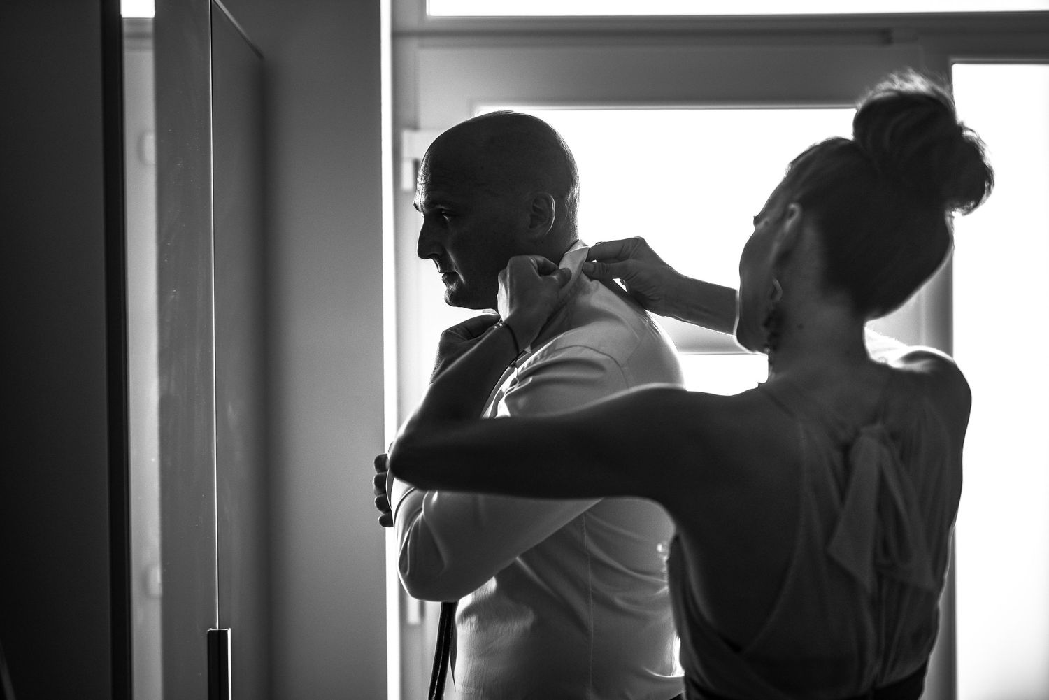 Black and white photo of a woman helping a man adjust his tie while getting ready for a wedding.