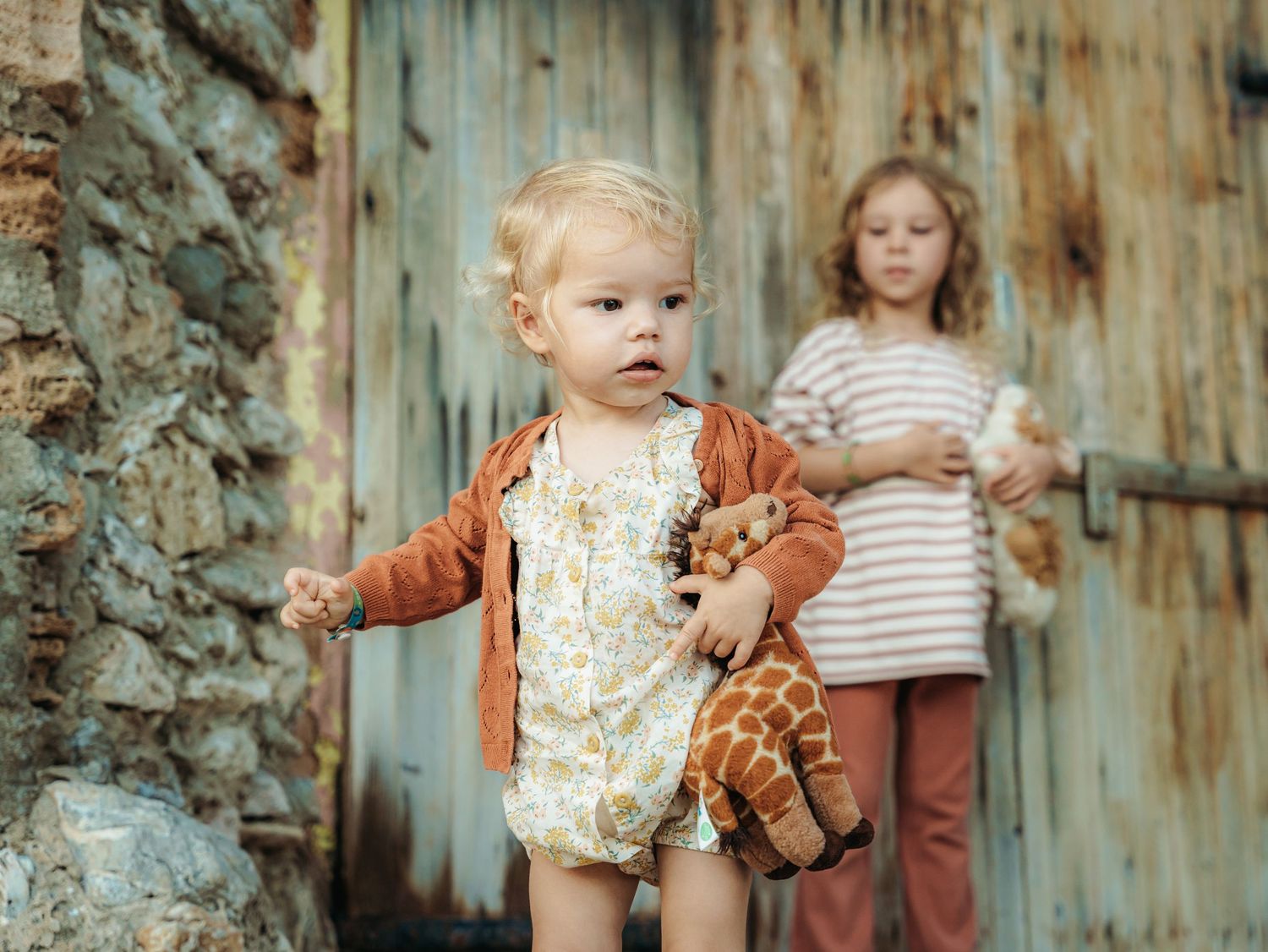 Toddler girl in floral romper holds stuffed giraffe outdoors near rustic stone and wood backdrop.