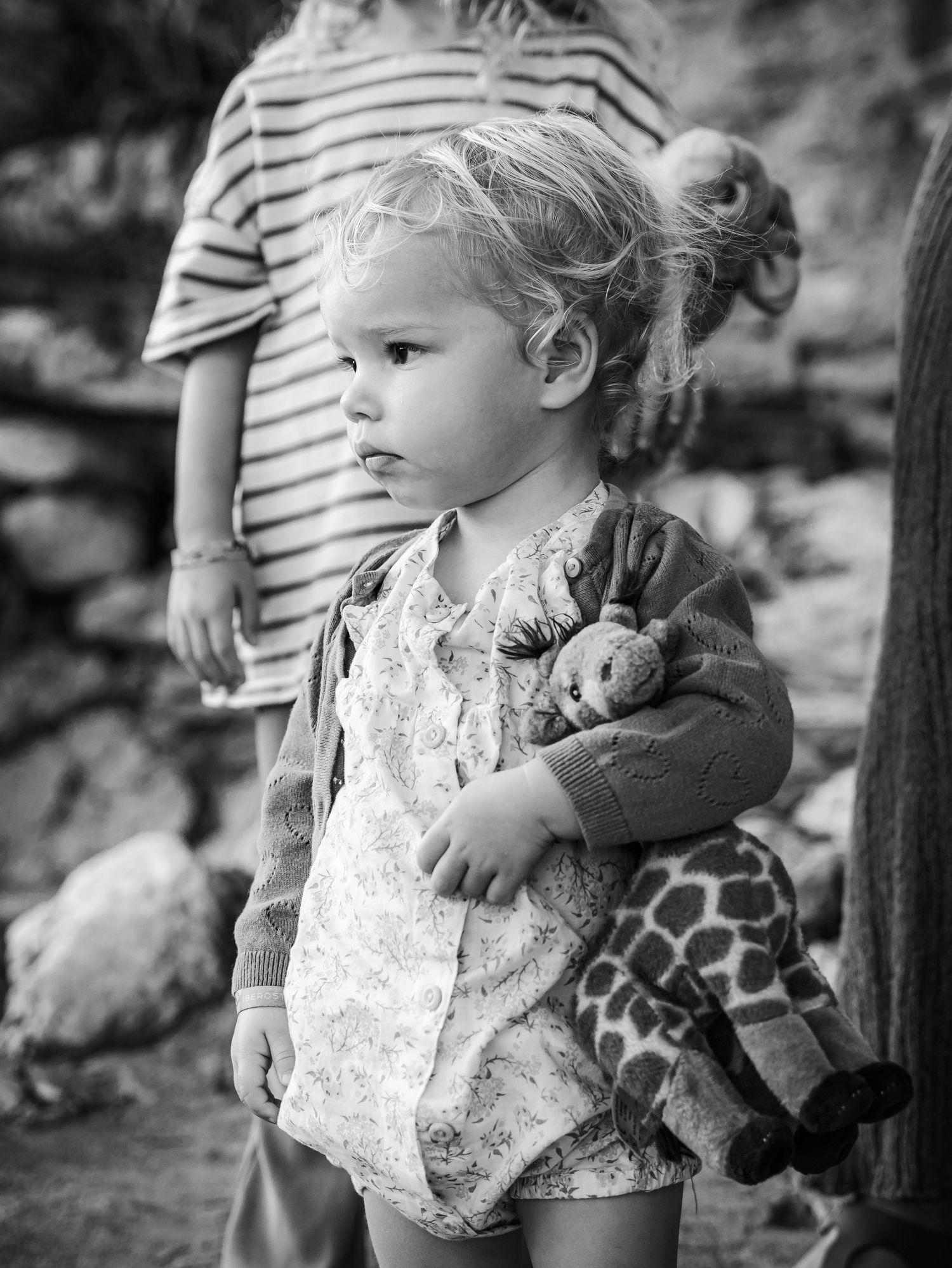 Black and white portrait of a toddler in a floral romper holding a giraffe stuffed animal outside.