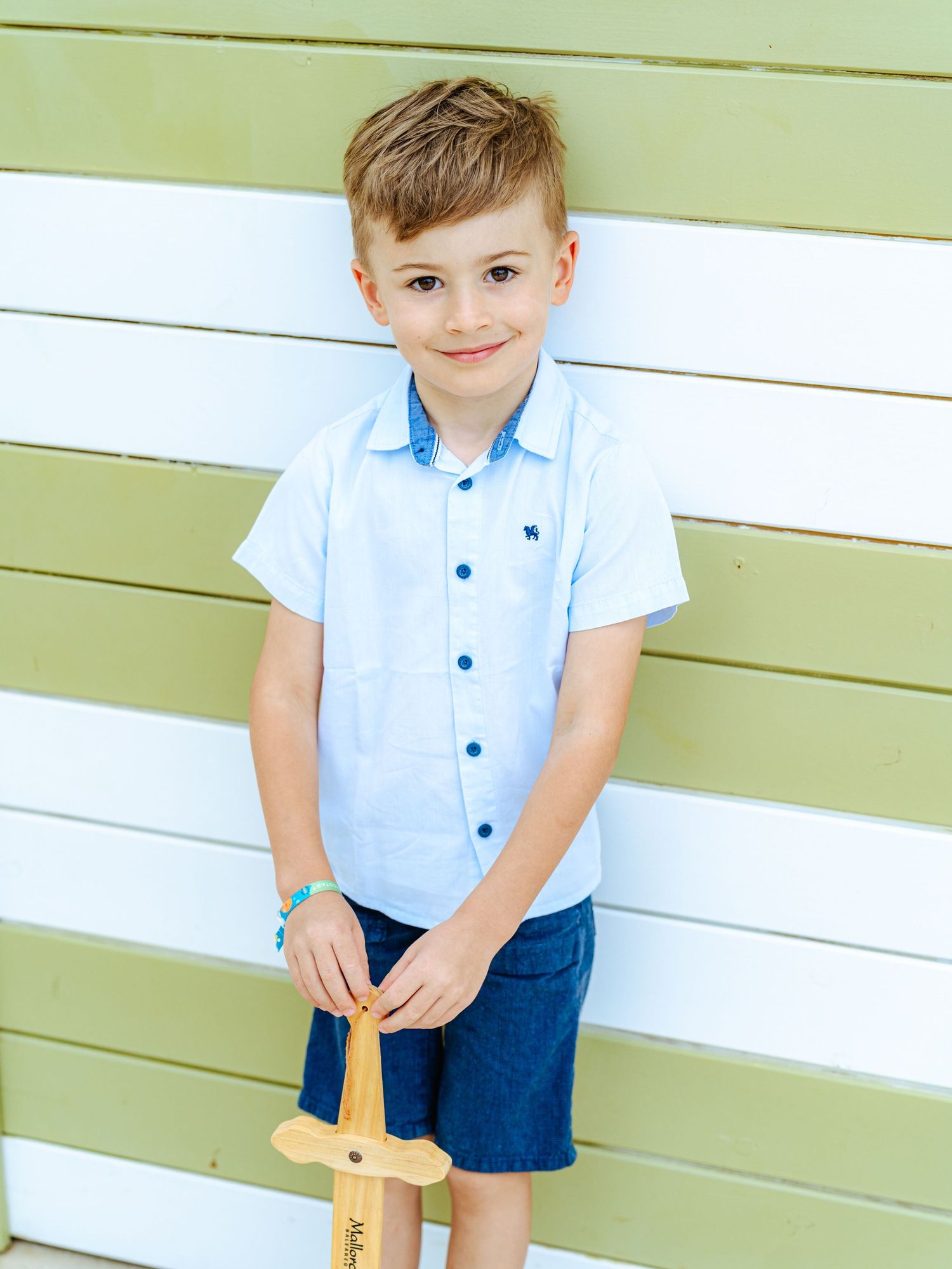 Young boy in light blue button shirt smiling while holding a wooden toy sword outdoors.