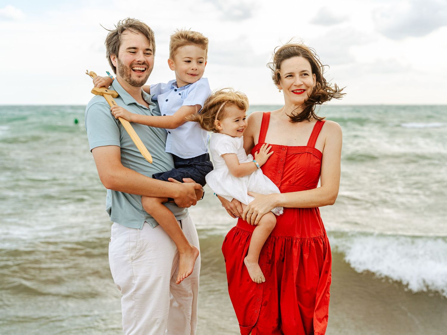 Smiling family of four at the beach, father and mother each holding a young child, ocean backdrop.