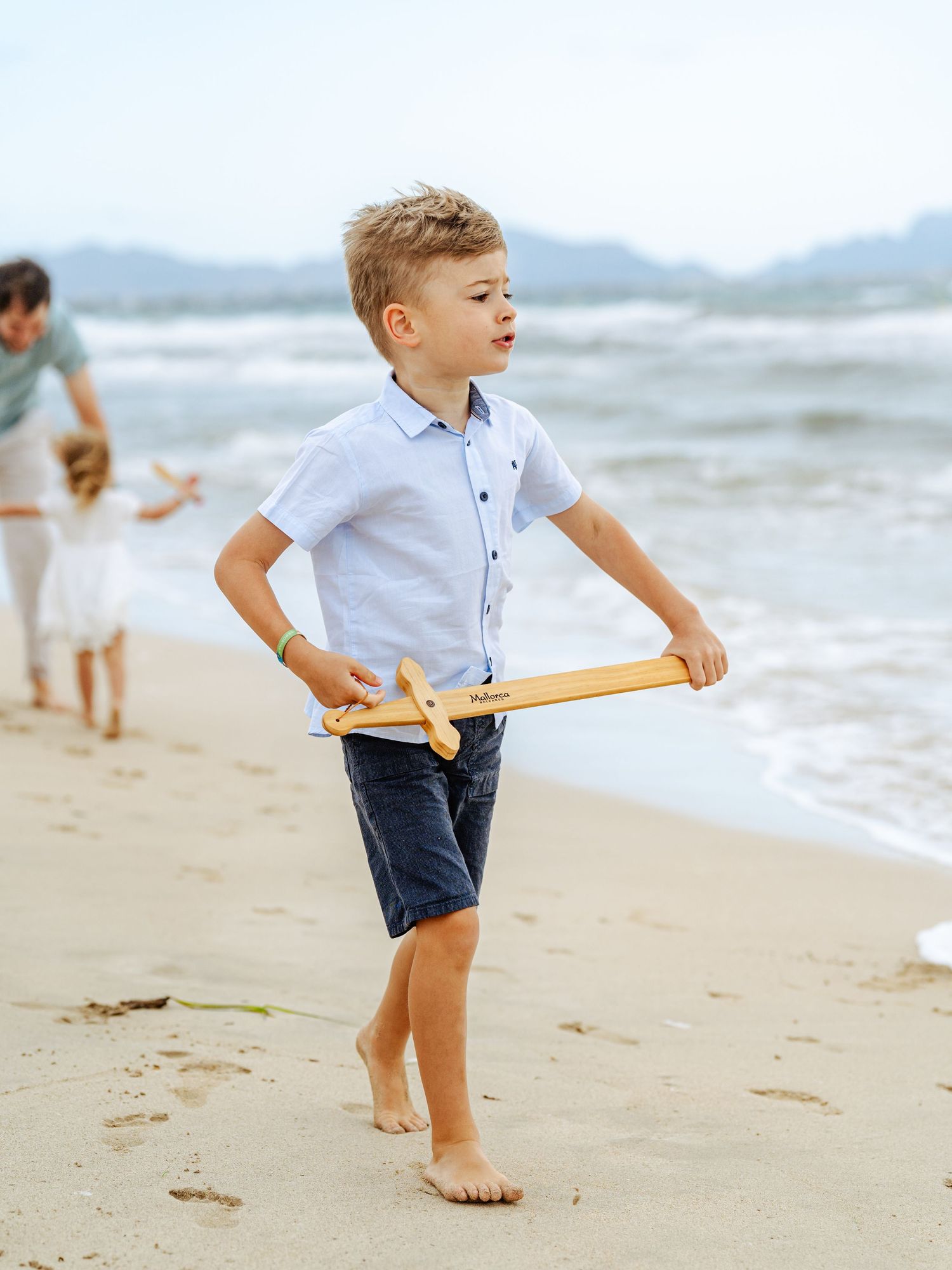 Boy in blue shirt and navy shorts holds wooden toy airplane on sandy beach.
