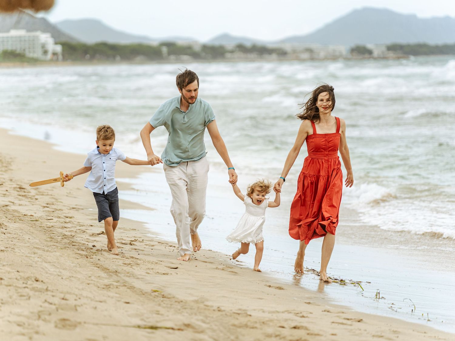 Happy family of four running along sandy beach with mountains in background.