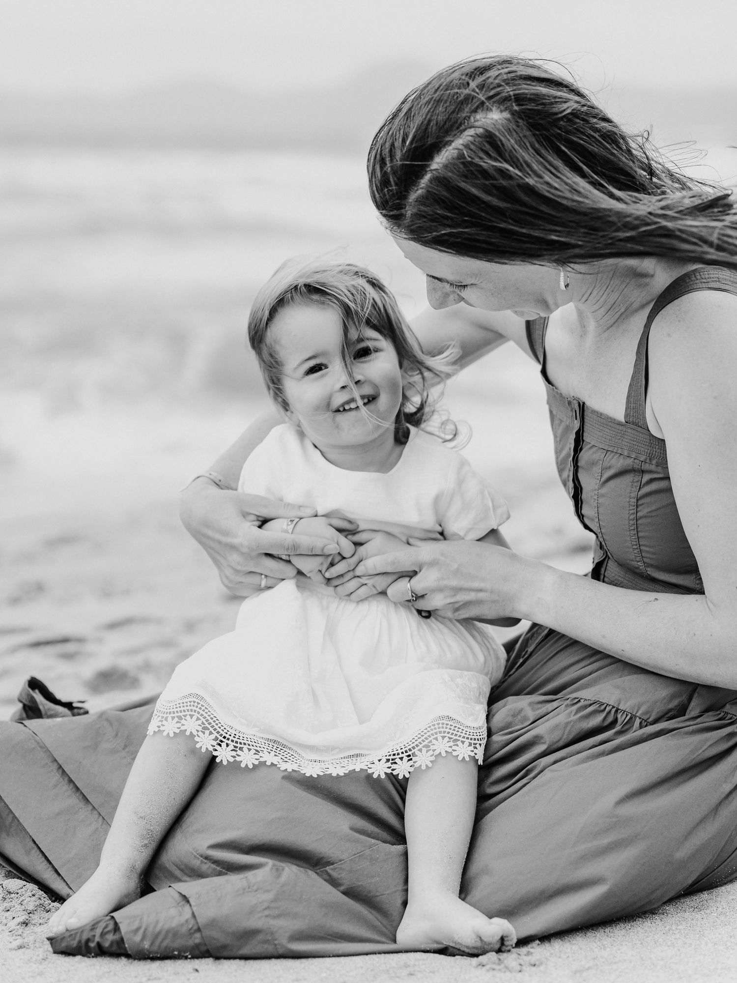 Black and white photo of laughing toddler girl sitting with mother on beach blanket.