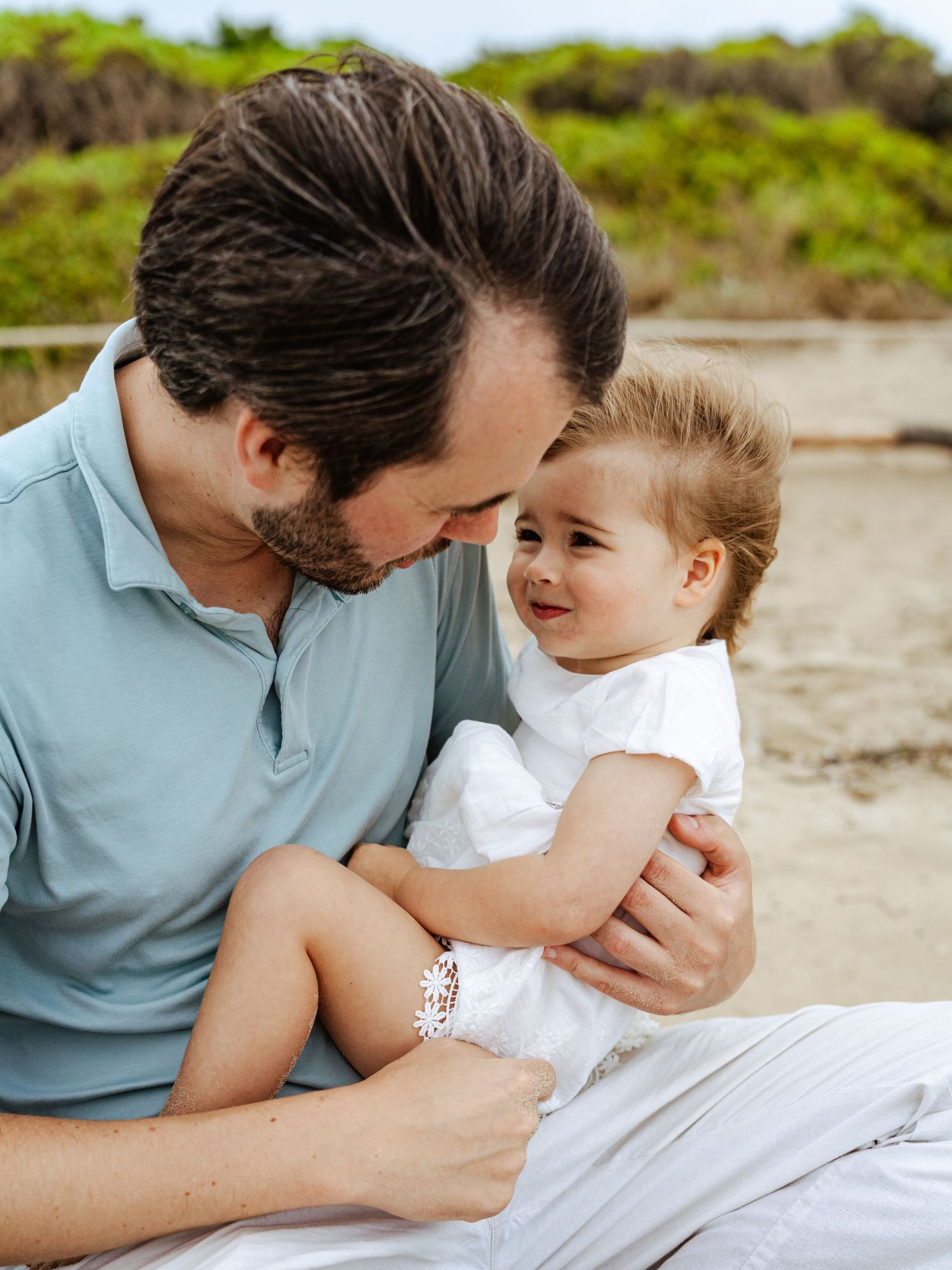 Loving father in light blue polo smiling at happy baby girl sitting on his lap at beach.