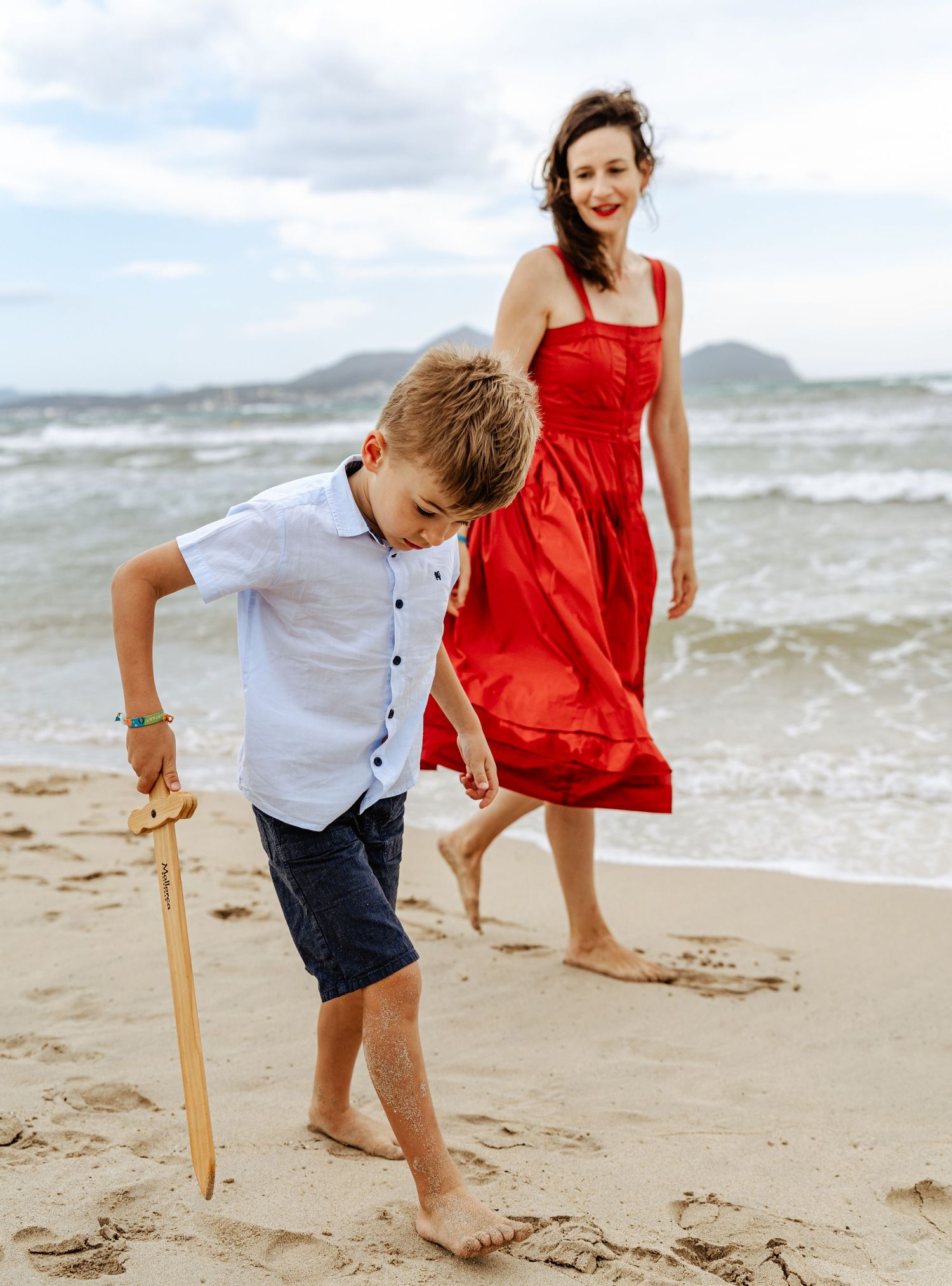 A young boy draws in the sand with a wooden stick as a smiling woman in a red dress walks behind him.