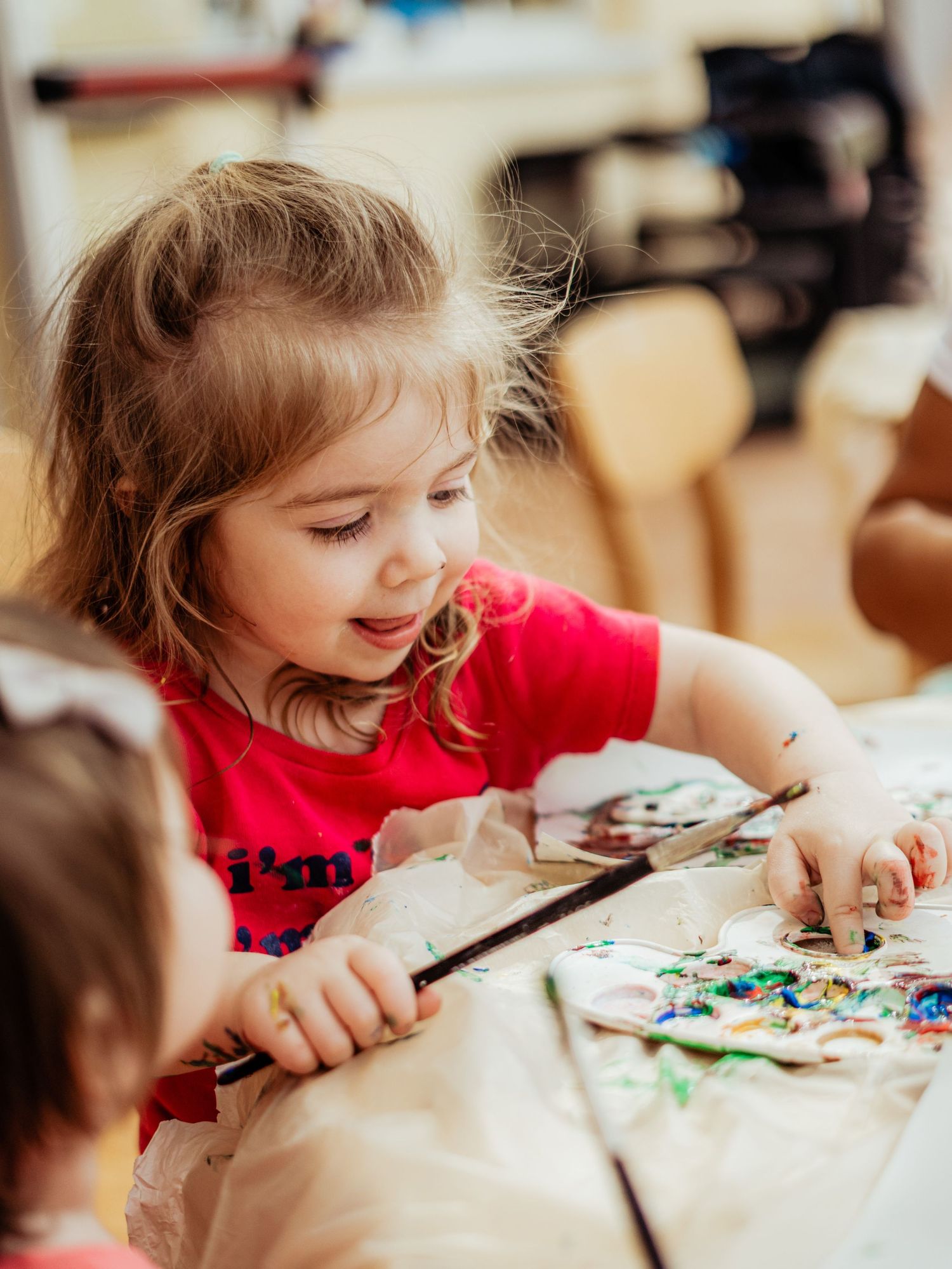 A young girl in a red shirt focuses intently on a painting activity at a wooden table.