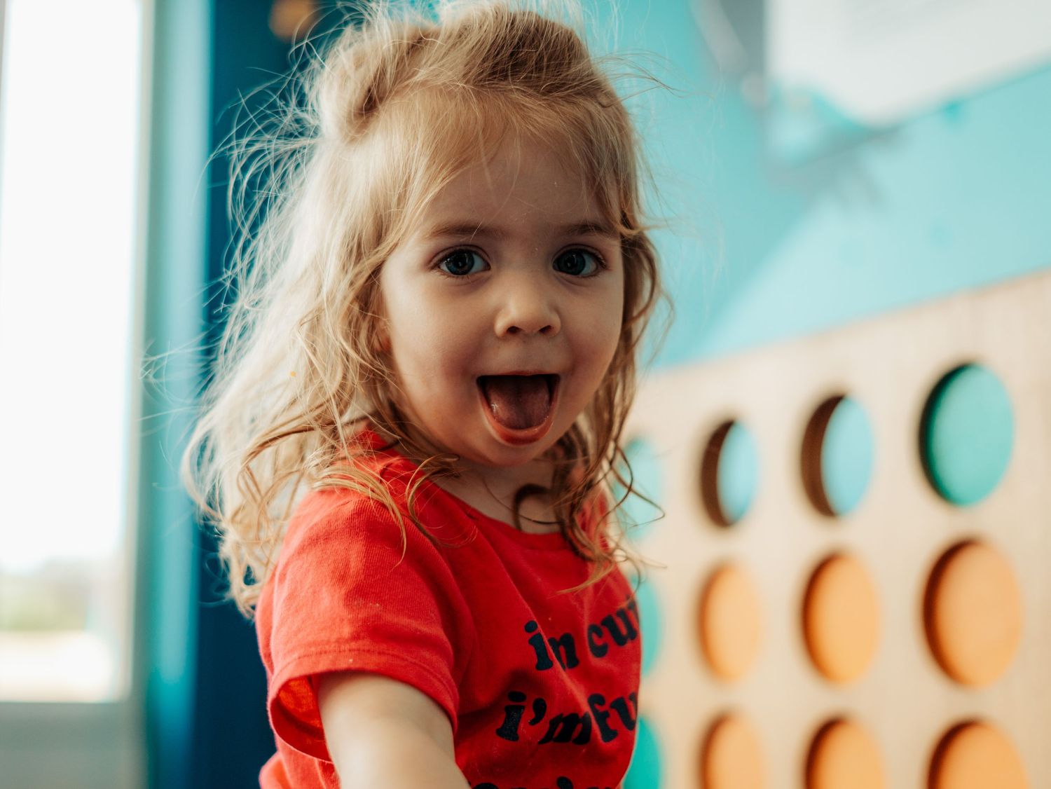 Excited toddler girl with blonde hair in red shirt playing near wooden toy wall with colorful circles.
