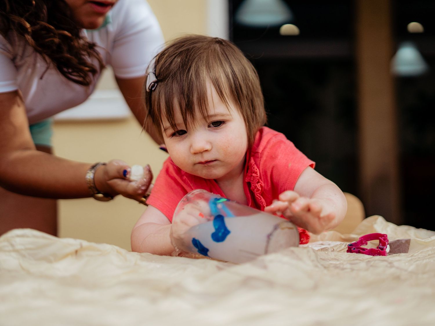 Toddler girl in coral shirt reaching across a table while an adult assists her with an activity.