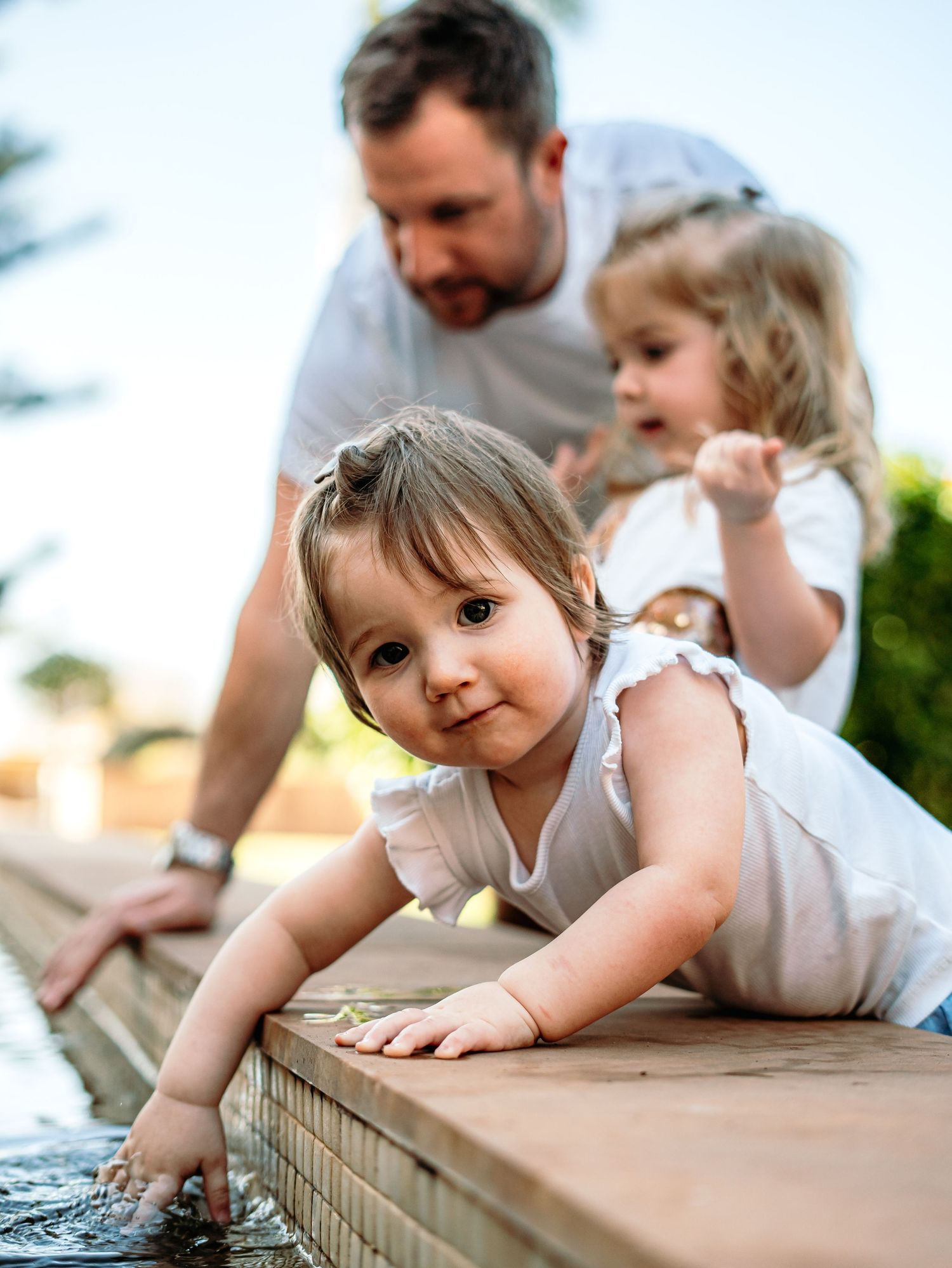 Father watches two young girls play at the edge of a reflective water feature in a sunny garden.