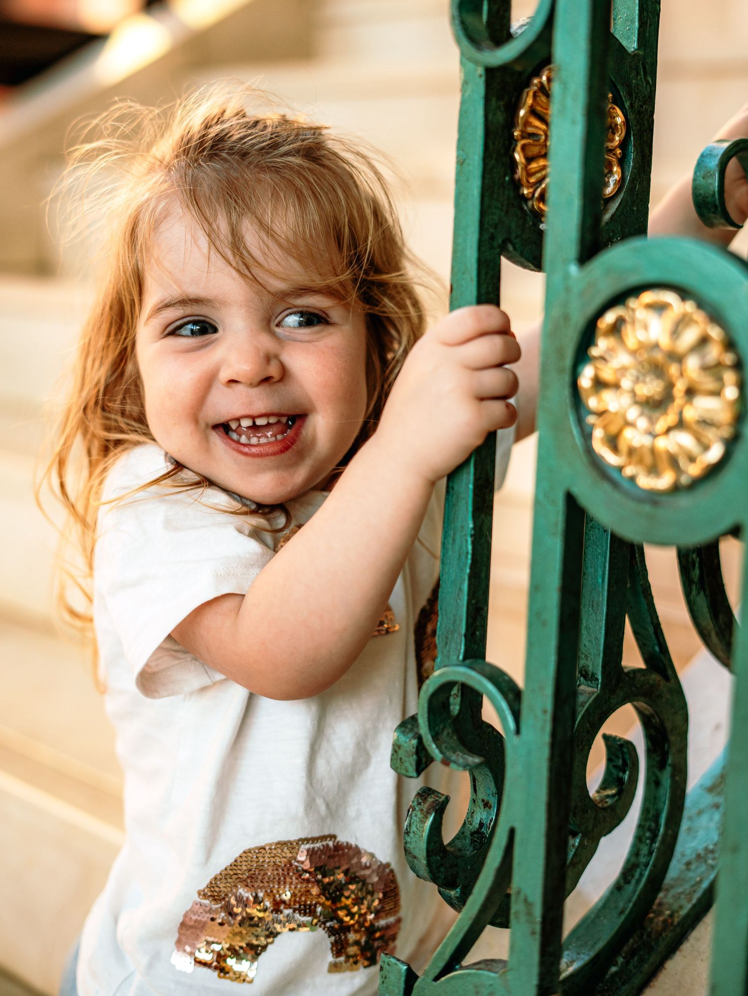 Smiling toddler girl with blonde hair holding green ornate iron gate outdoors.