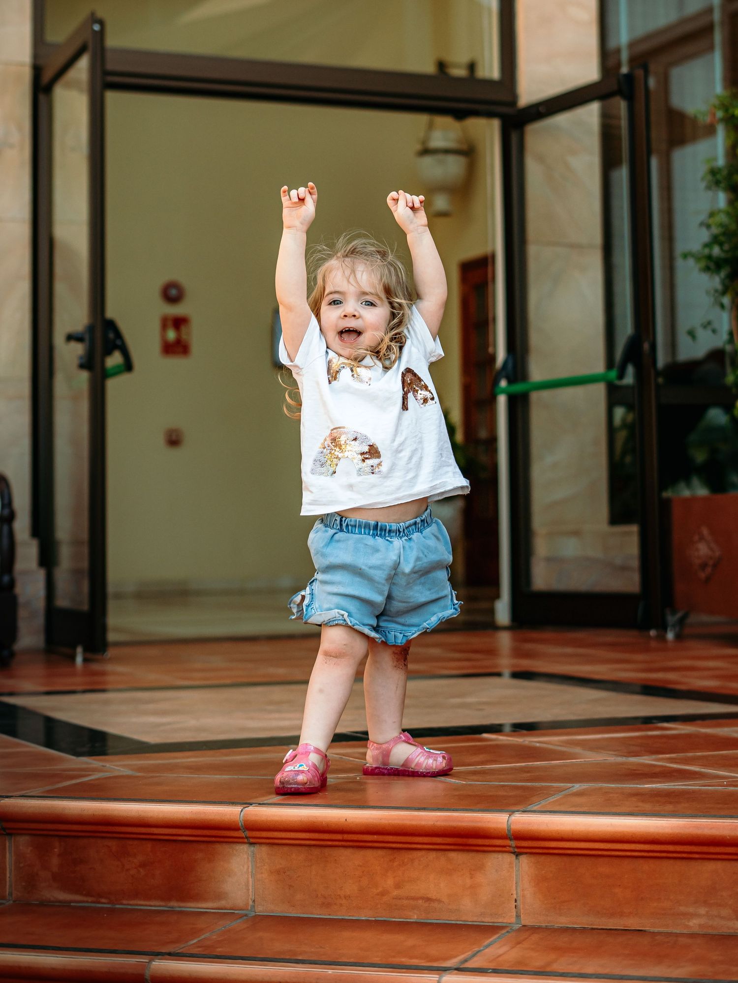 Happy toddler girl in white tee and denim shorts cheering with arms raised at hotel entrance.