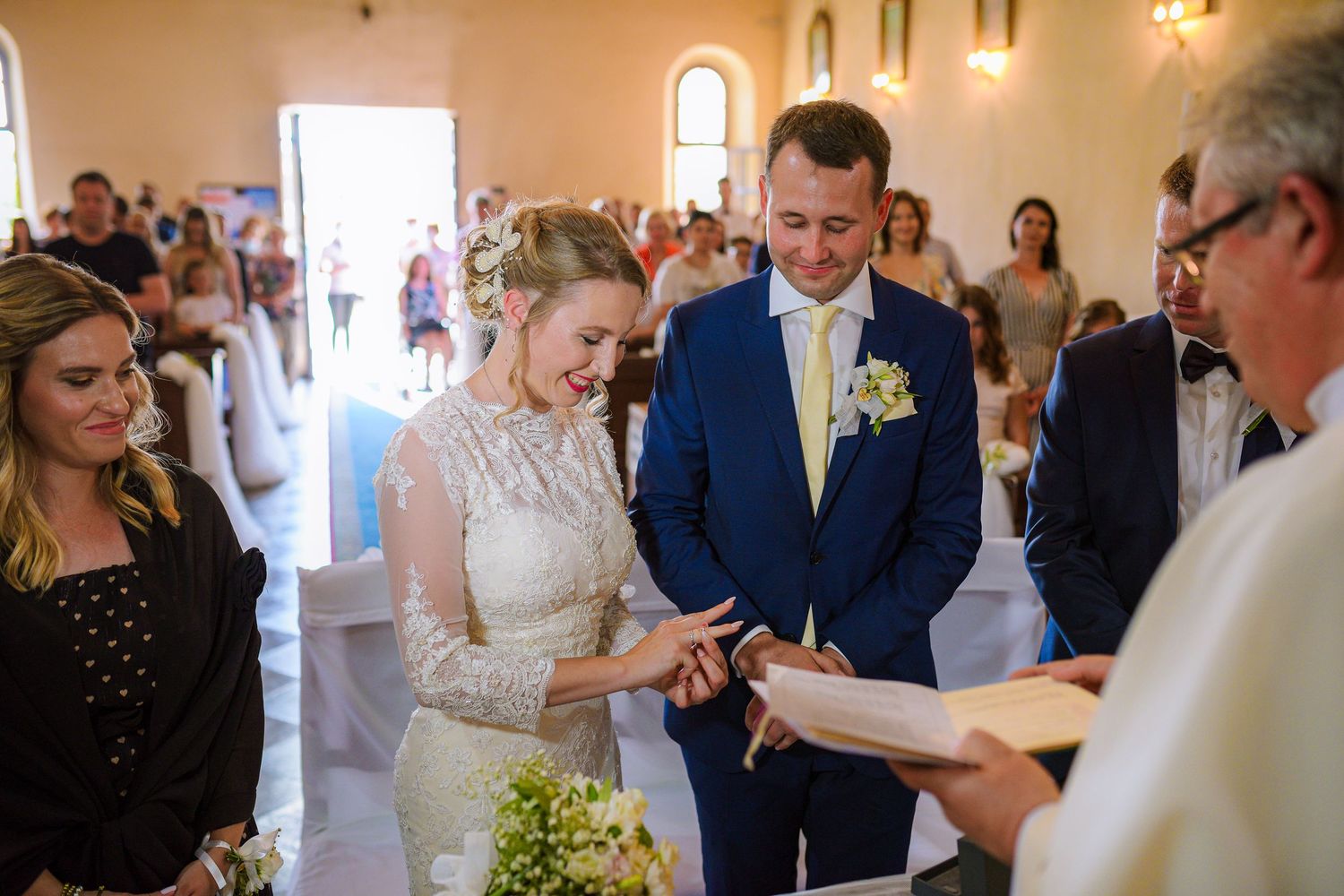 Smiling bride placing wedding ring on groom's finger during church ceremony with guests watching.